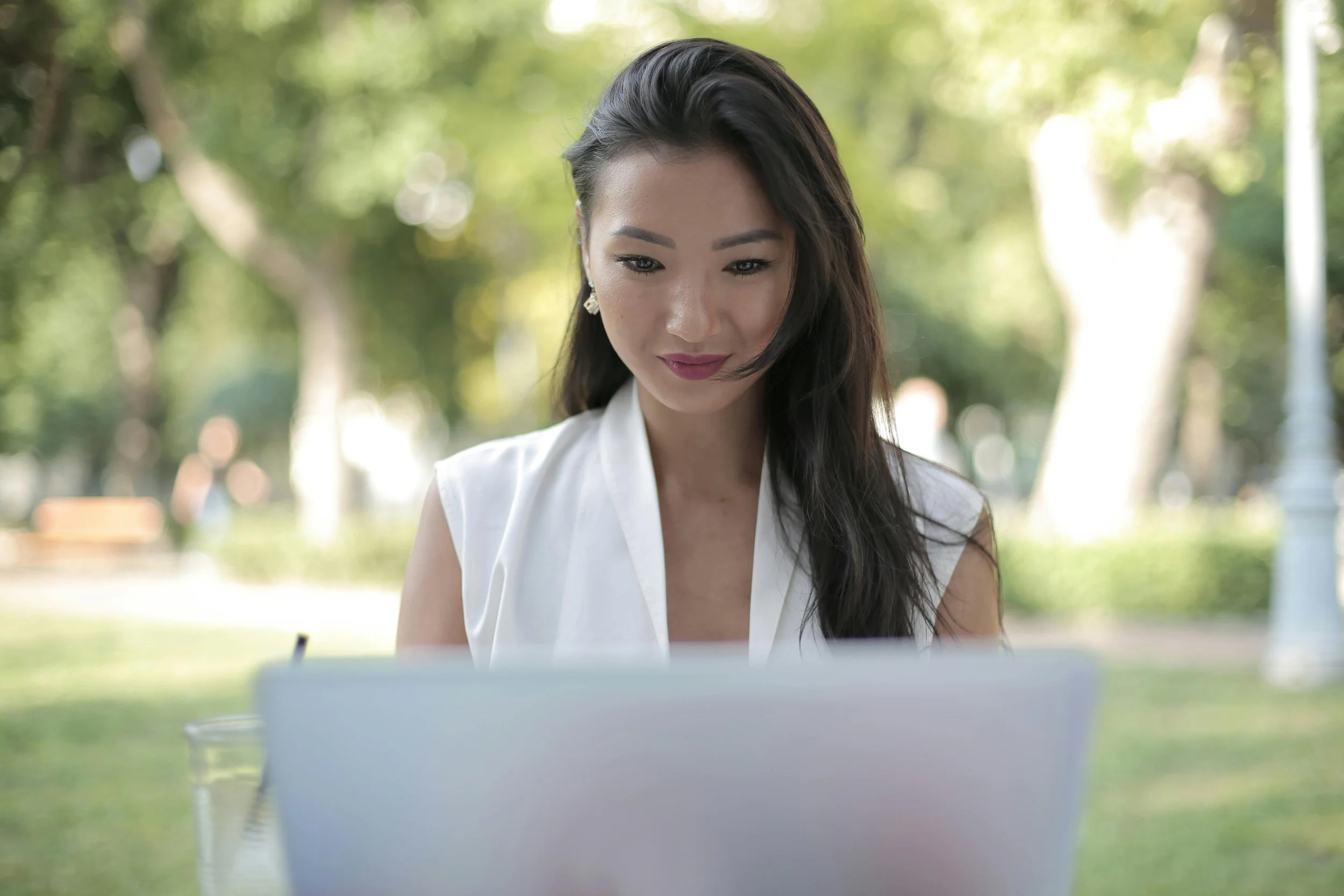 A woman with long dark hair and earrings sitting outdoors, looking at a laptop. The background shows a green, blurred park or garden scene.