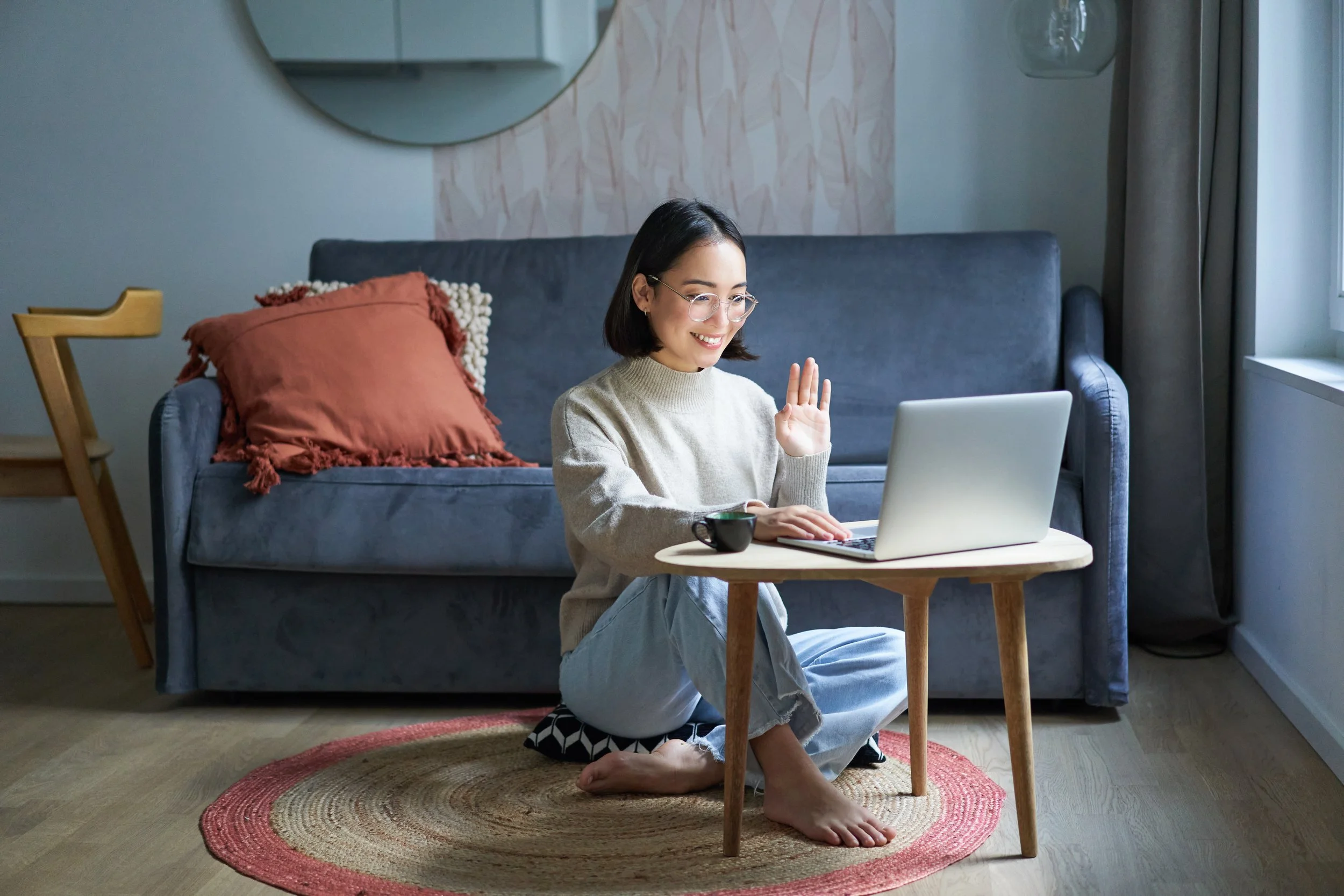 A woman with glasses, sitting on a rug on the floor, waving and smiling at her laptop on a small wooden table. She is in a cozy living room with a gray sofa, a round mirror on the wall, a red cushion, and a window with curtains.