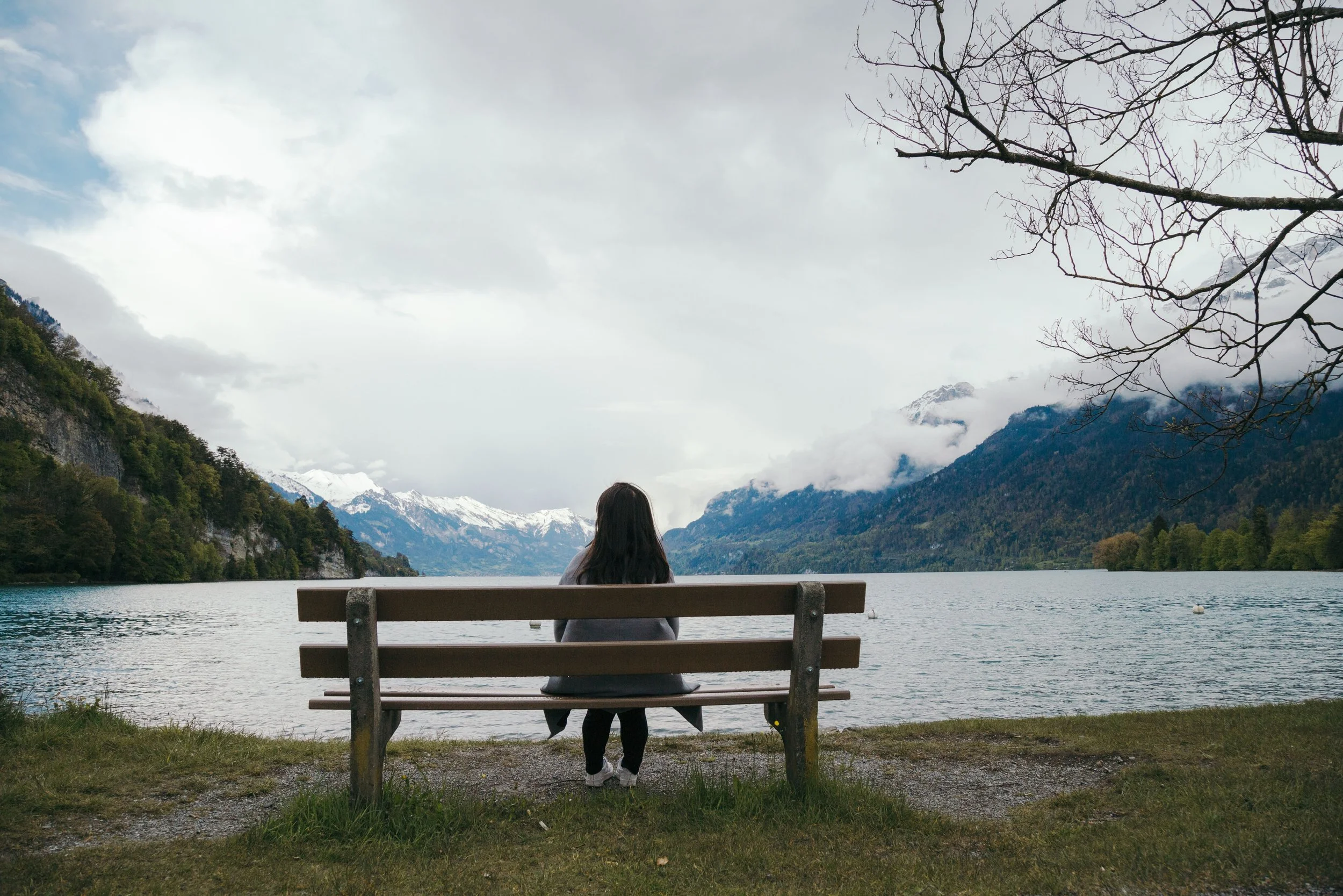 A person with long dark hair sitting on a bench by a lake, facing mountains and snow-capped peaks, with trees and cloudy sky in the background.