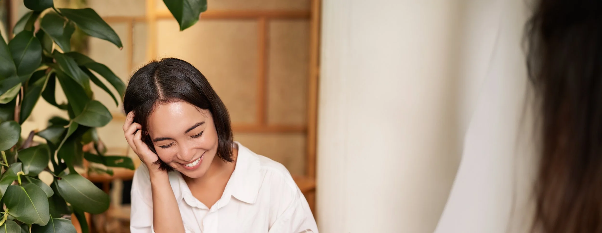 A woman with short dark hair smiling and touching her ear, sitting next to a large leafy green plant in an indoor setting.