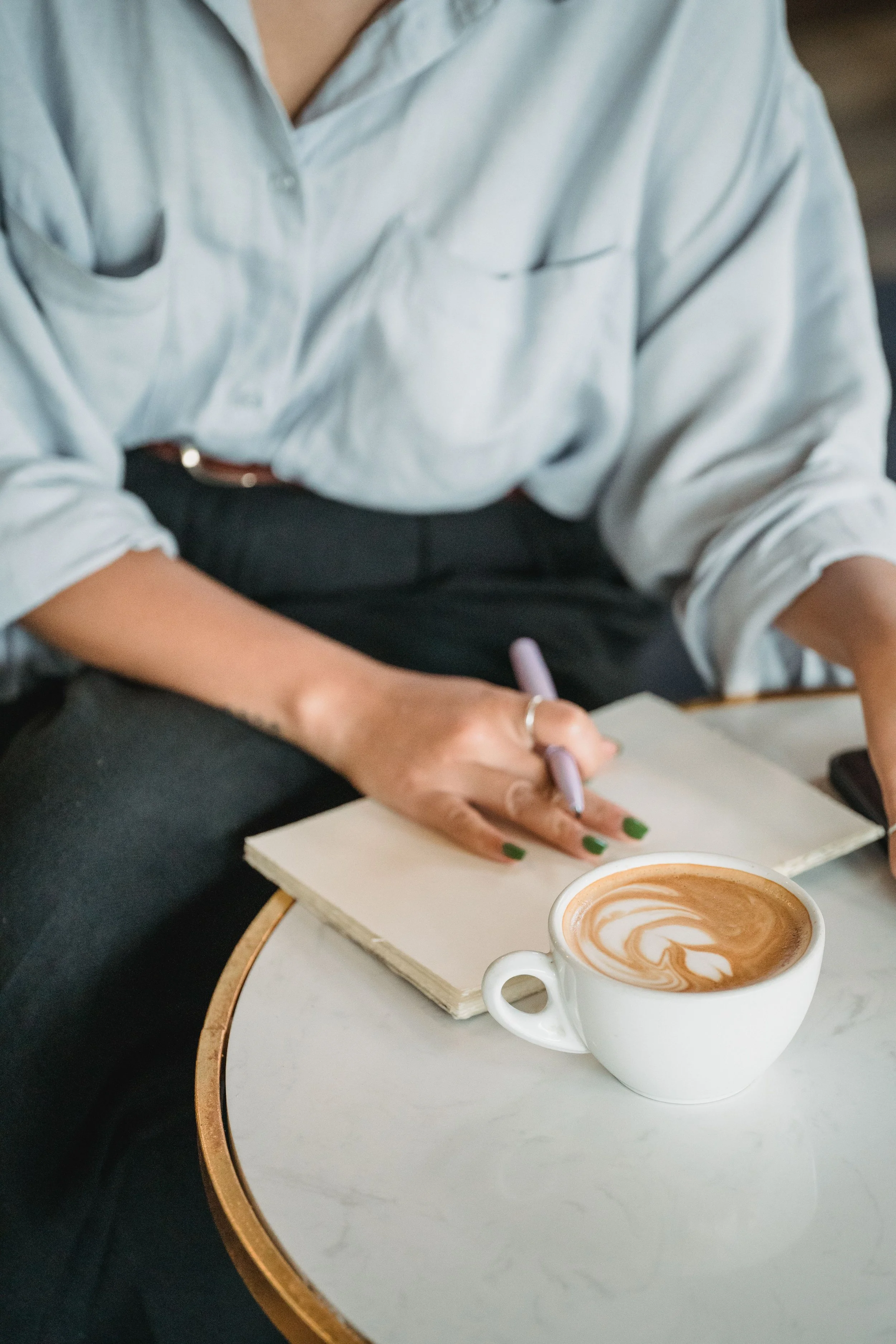 A person with green nail polish sitting at a table with a cup of coffee that has latte art and a notebook, writing with a pen.