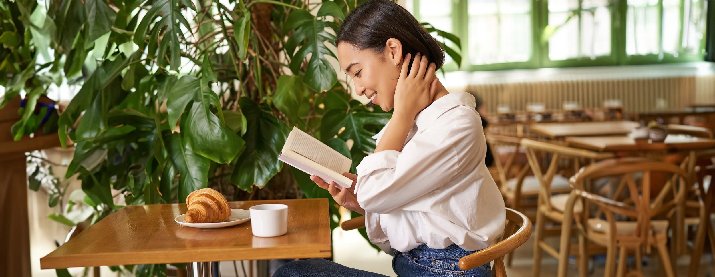 A young woman with dark hair, wearing a white shirt, sitting at a wooden table in a cafe, looking at a book with a smile. There is a croissant on a plate and a cup of coffee on the table. Green plants and wooden furniture are in the background.
