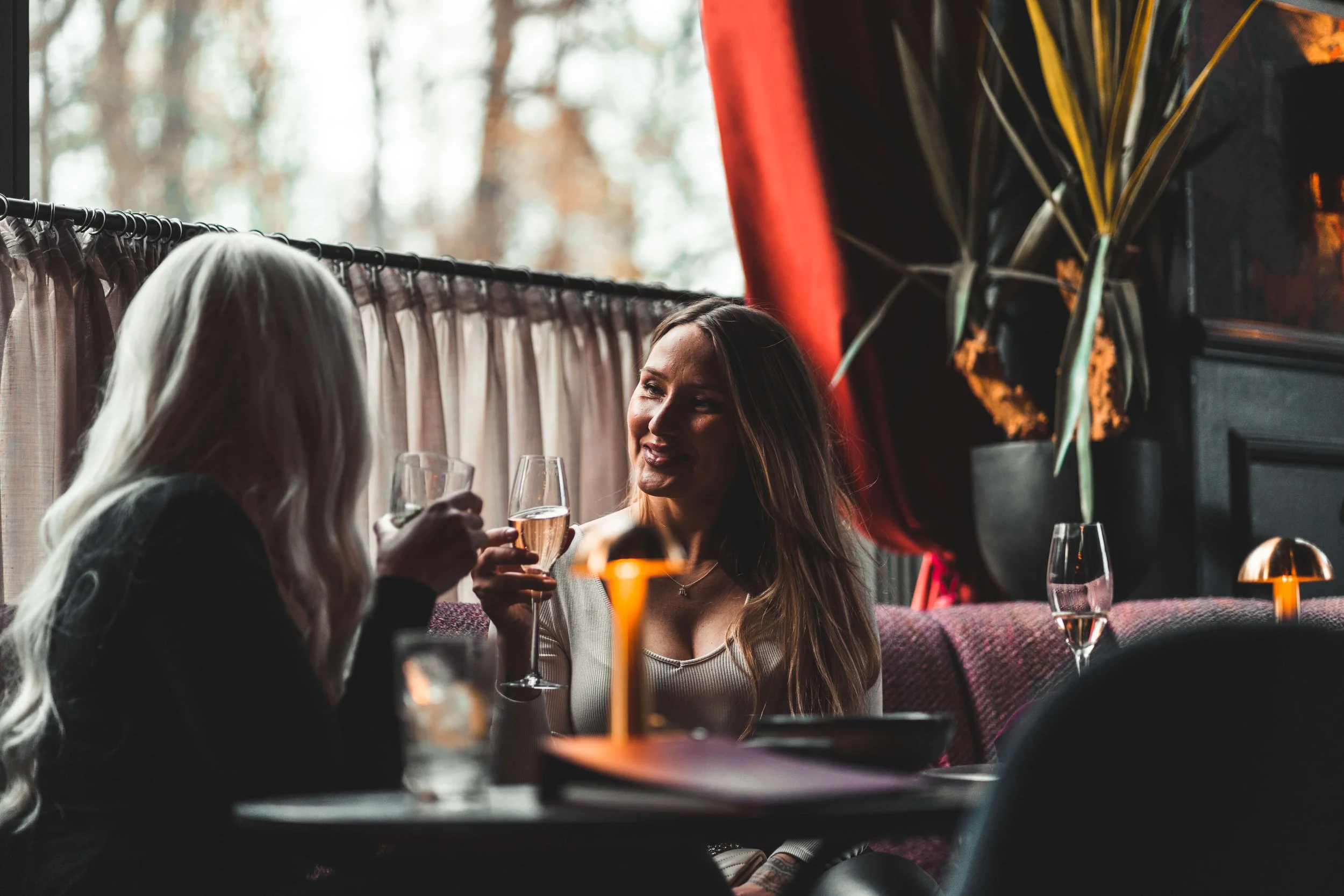 To kvinner sitter i en bar eller restaurant, smiler og skåler med vinglass. Stemningen er koselig og avslappet, med dempet belysning og dekorative elementer som planter og gardiner.