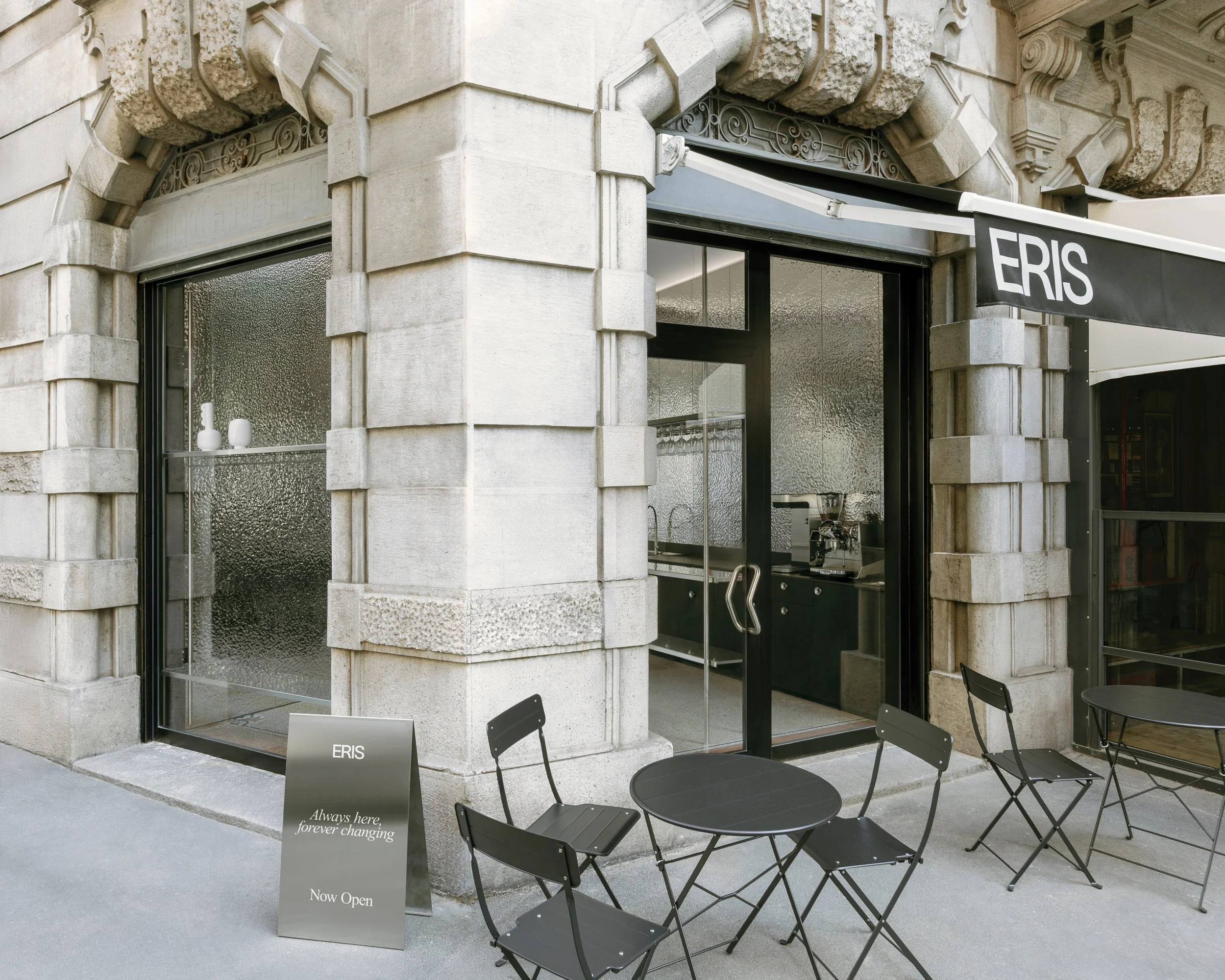 Esterno di un negozio con ingresso vetrato, decorazioni in pietra e un'insegna nera con la scritta 'ERIS'. Davanti ci sono tavoli e sedie nere con adesivi. Un cartello bianco con scritto 'Always, here forever changing' e 'Now Open' in inglese.