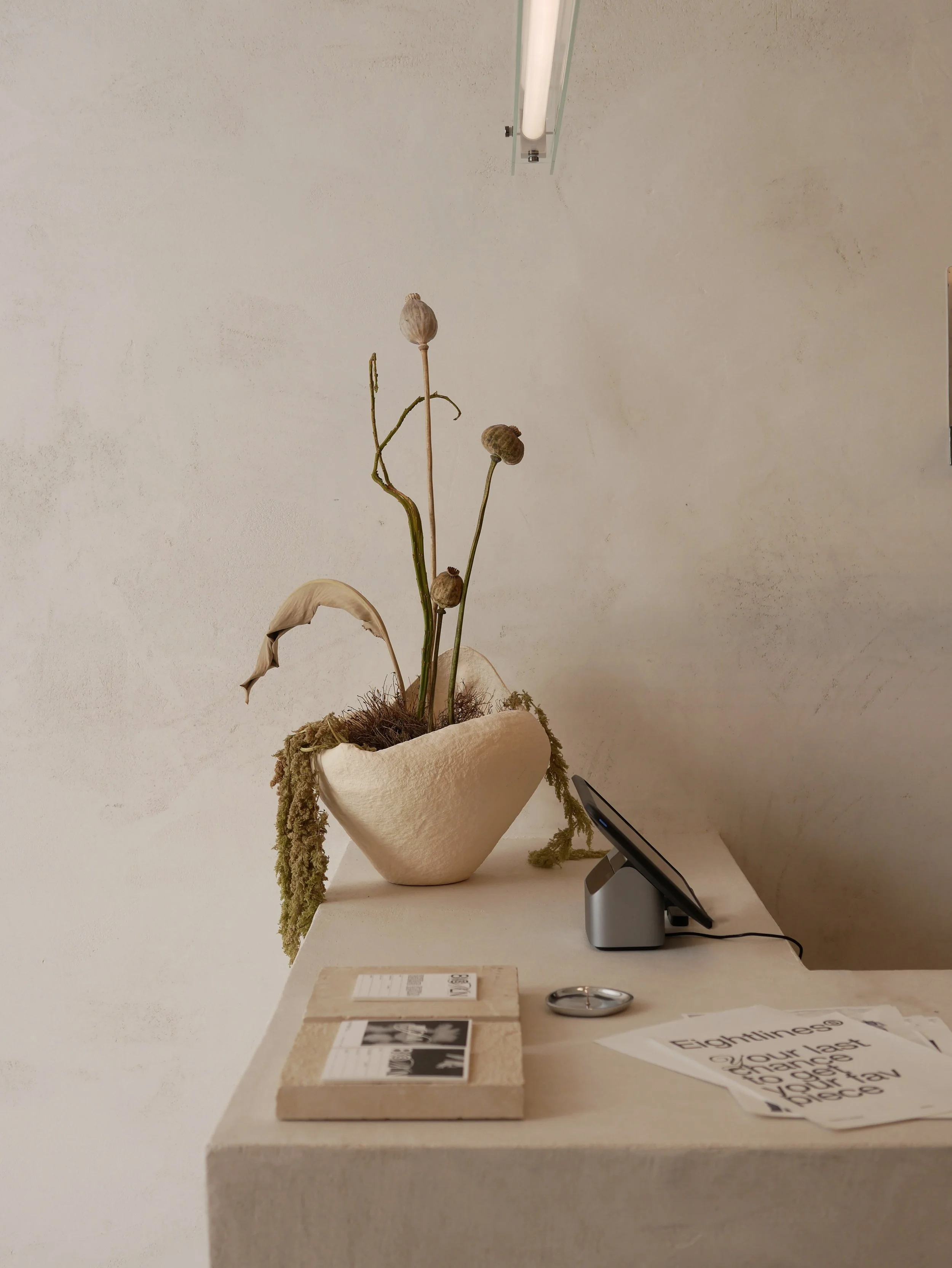 Vasetto d'arte con piante secche al centro, posta su un tavolo bianco con vari oggetti tra cui brochure, un telefono e un orologio. Sullo sfondo, parete bianca e lampada sopra