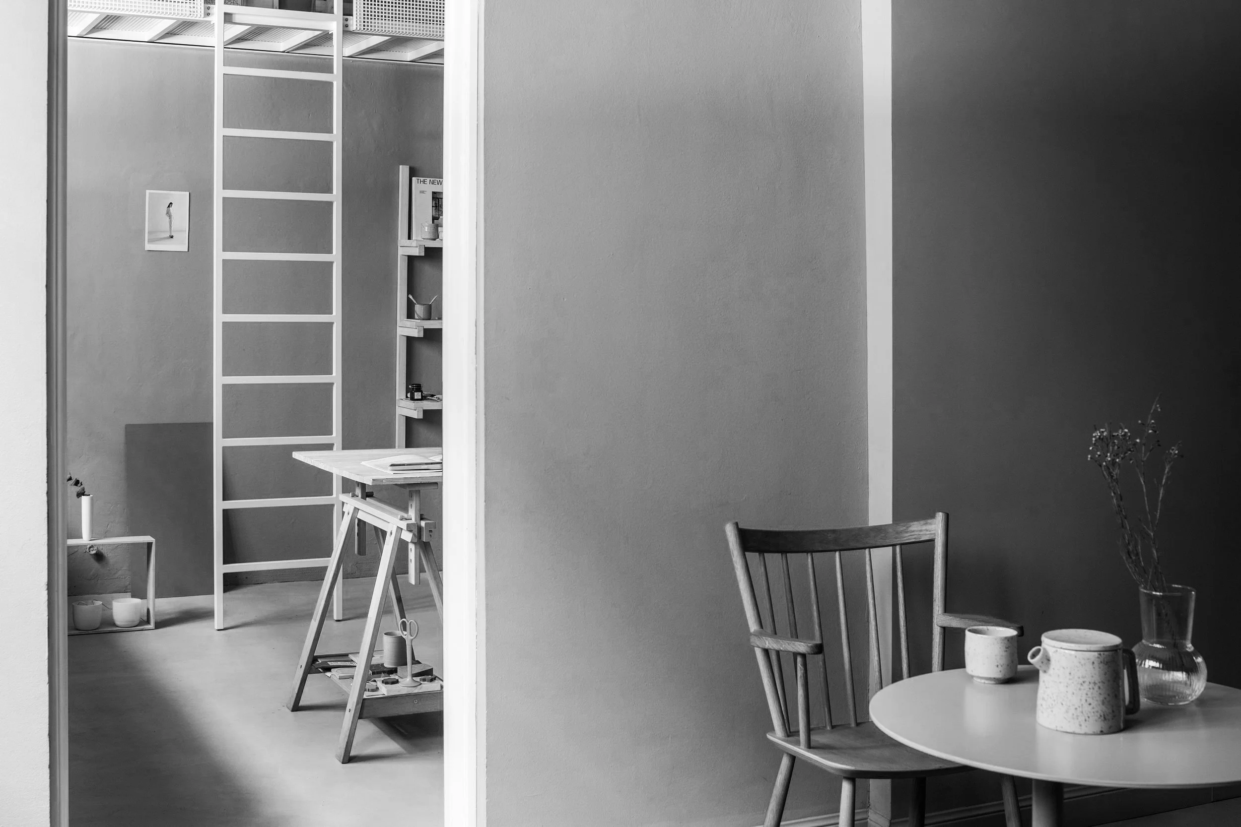 Interno di un'abitazione in bianco e nero con un tavolo rotondo, due sedie e vasi decorativi, e una parete che separa la stanza da un'area con scaffali e libri.