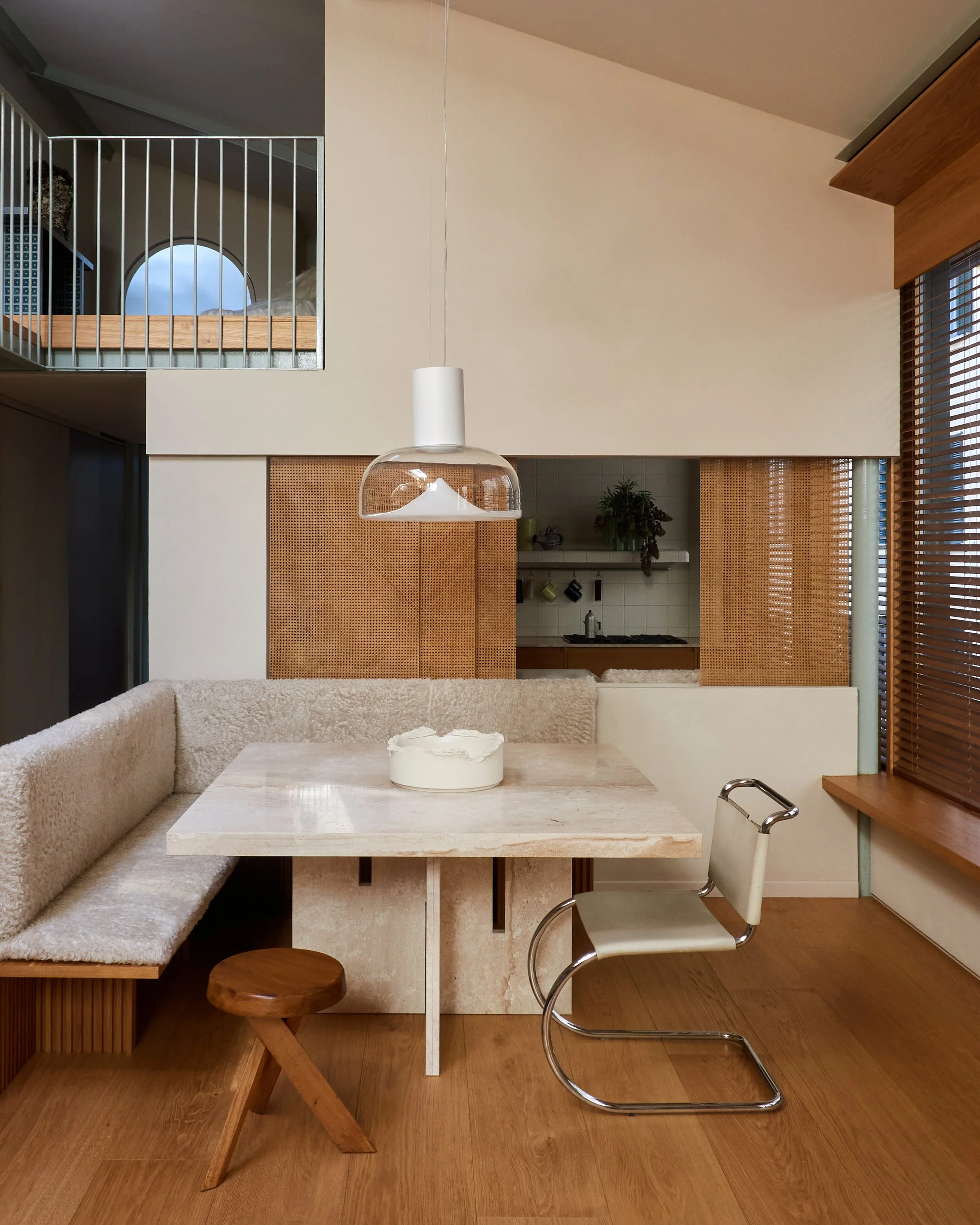 Interno di una cucina moderna con un tavolo in marmo, un divano in tessuto beige, una sedia in metallo e legno, lampada a plafon e finestre con tapparelle di legno. In alto, un soppalco con sbarre in metallo e alcune scatole e oggetti. Cucina sullo sfondo con piante, stoviglie e forno.