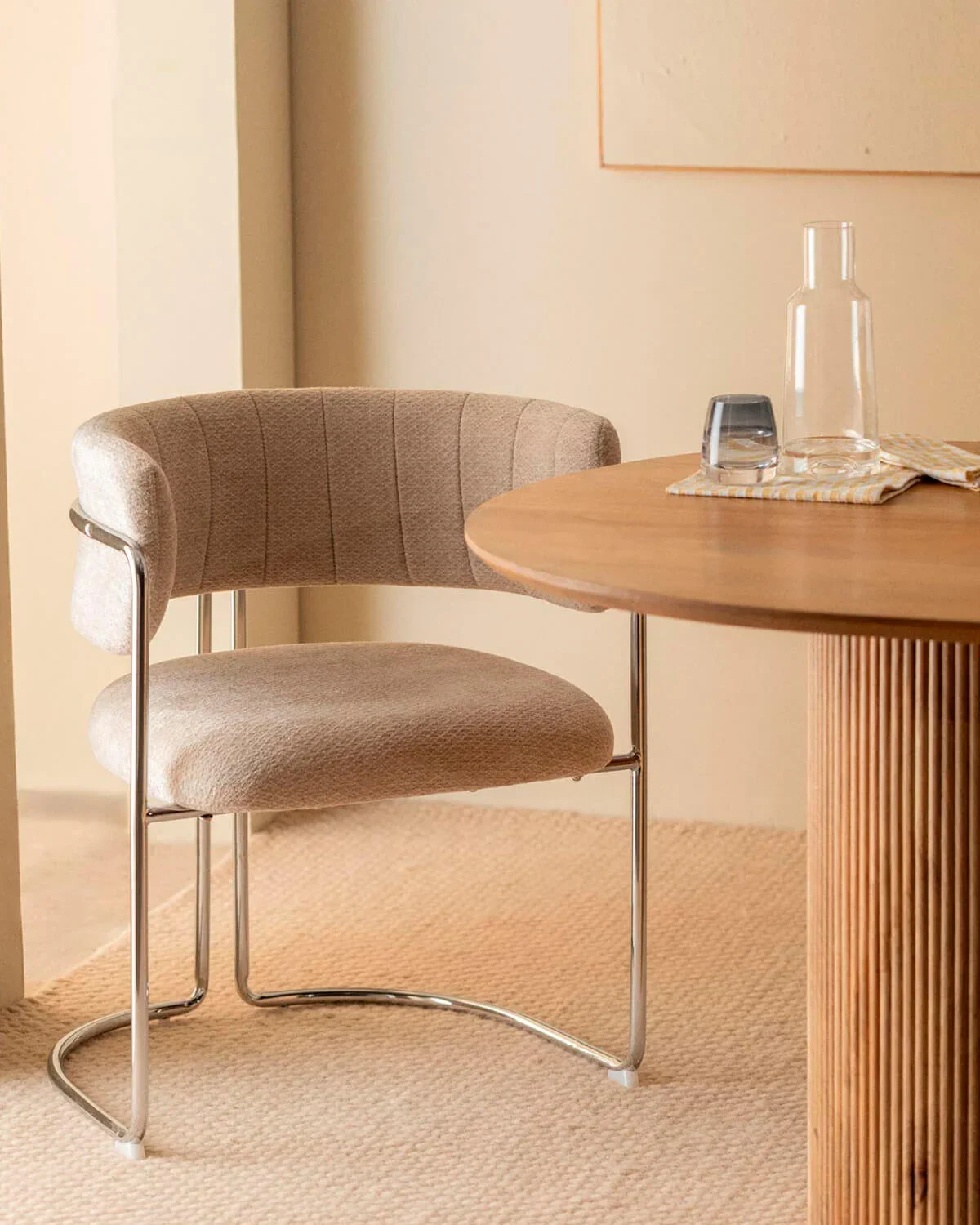 Sedia beige con struttura in metallo accanto a un tavolo di legno, con un piattino, due bicchieri e una bottiglia di vetro trasparente