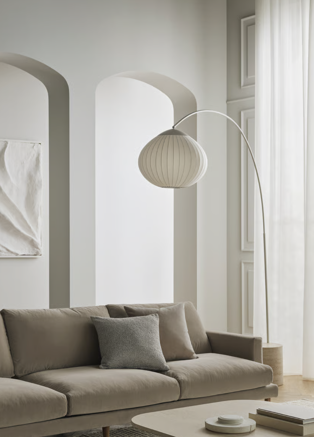Soggiorno minimalista con divano beige, cuscino grigio, lampada da terra bianca con arco e lampada da soffitto sospesa bianca, e finestre con tende leggere.