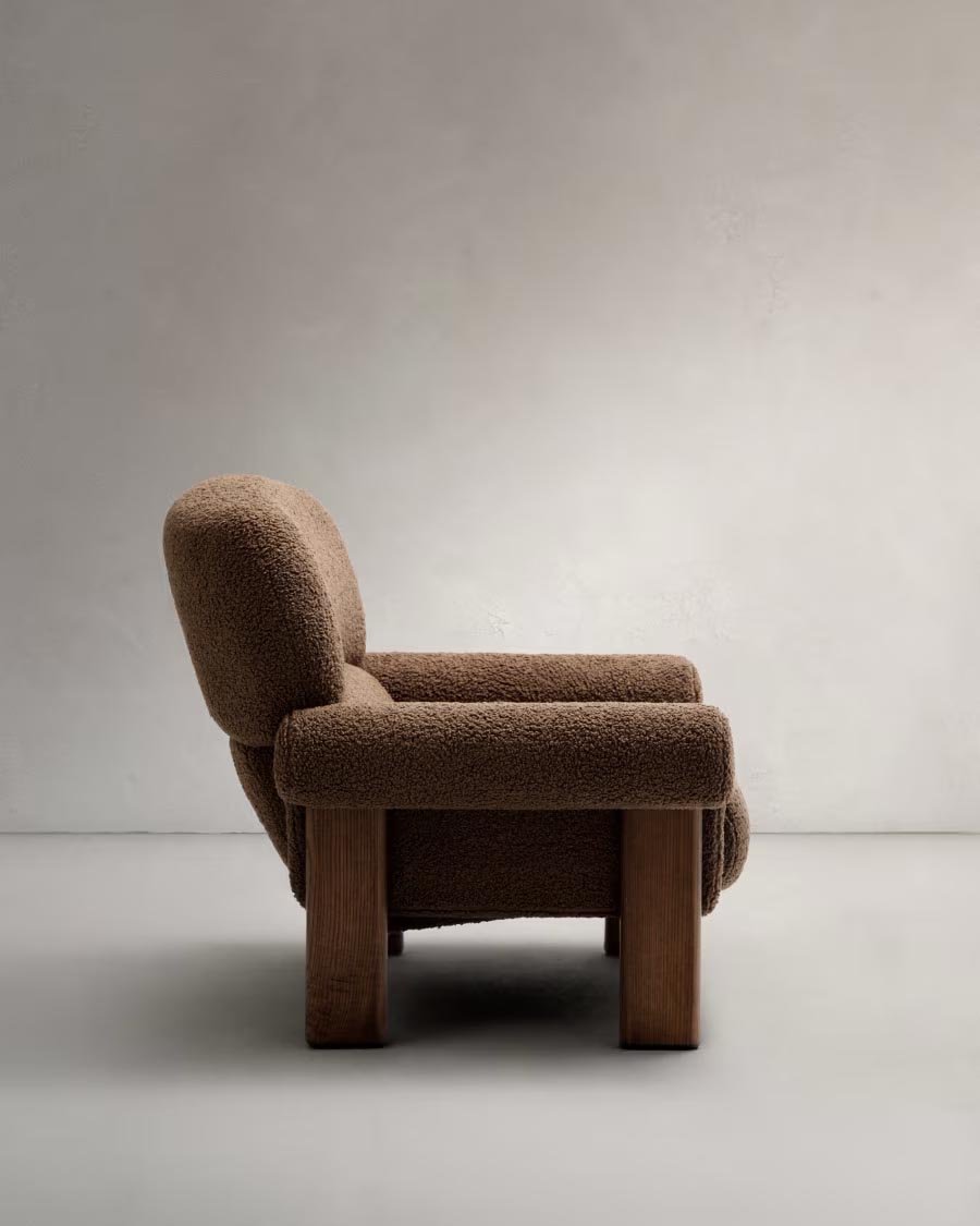 Poltrona imbottita in tessuto marrone con piedi in legno, vista laterale, in uno sfondo neutro.