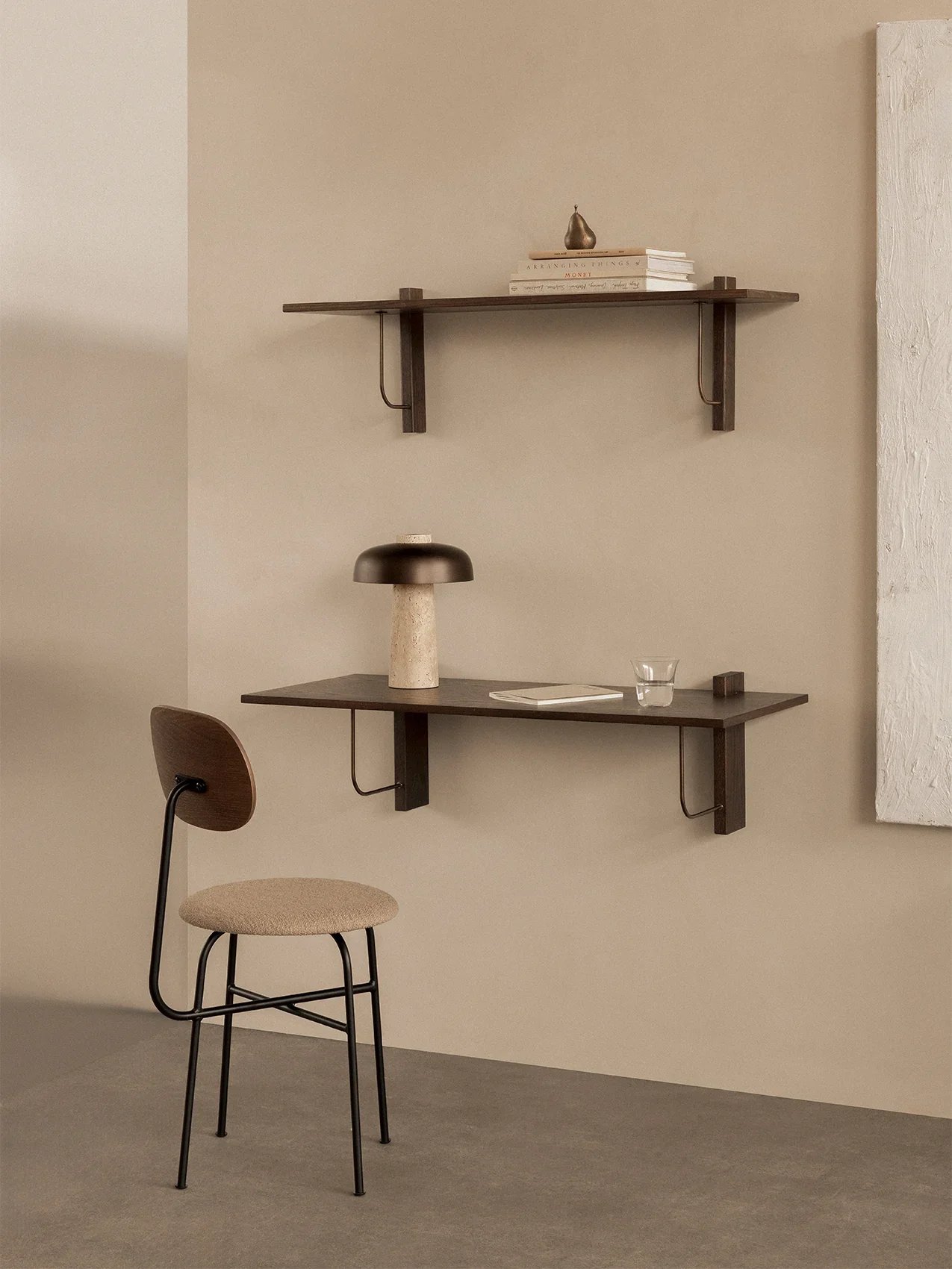 Interno minimalista con due scaffali di legno marrone scuro sulla parete beige, una sedia con strutture nere, seduta e schienale in tessuto beige, una lampada śtile da tavolo con base in pietra e paralume nero, un bicchiere e un libro sul tavolo.