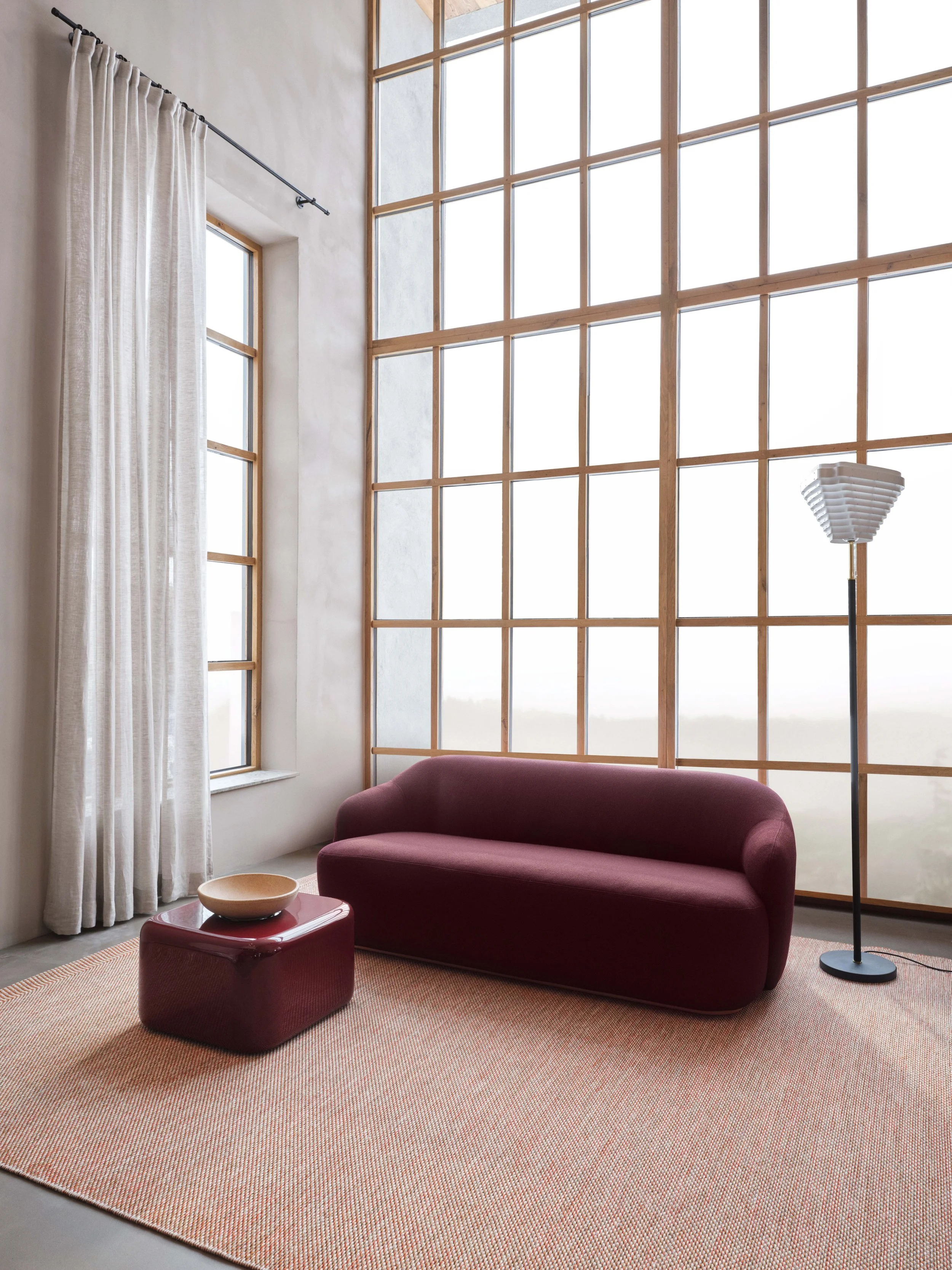Interno con grandi vetrate in legno, divano rosso, lampada da terra nera, tavolino bagliore rosso, tappeto beige e tende bianche in stoffa
