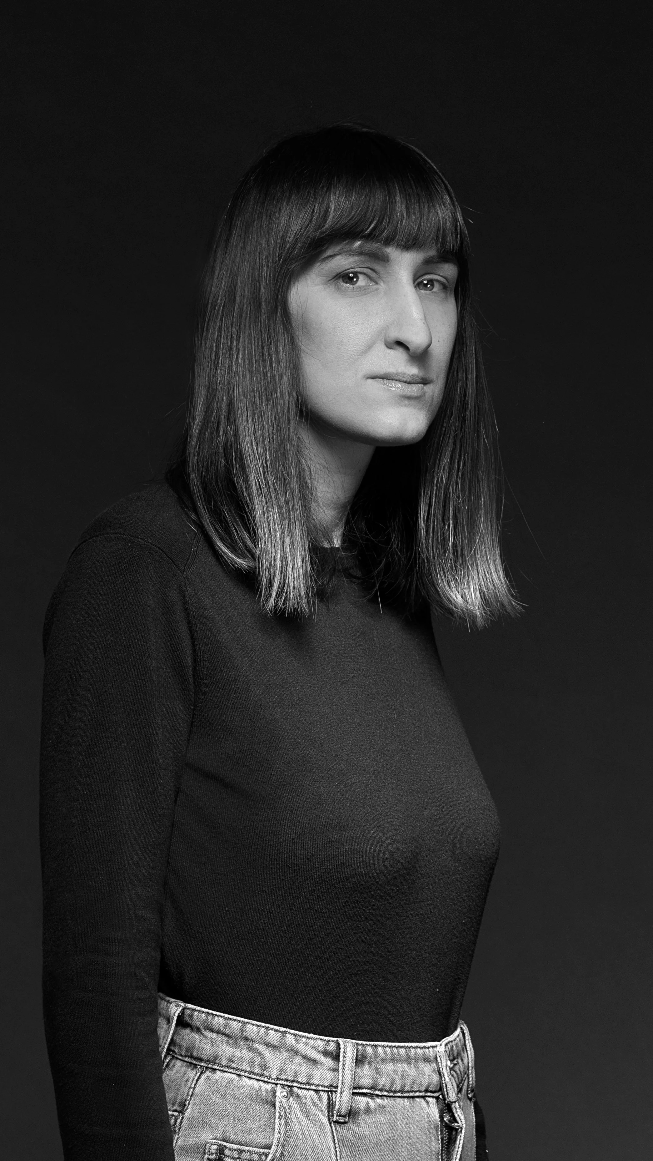 Ritaglio di una donna con capelli lunghi e lisci, indossa una maglietta nera e jeans chiari, posa contro sfondo nero.