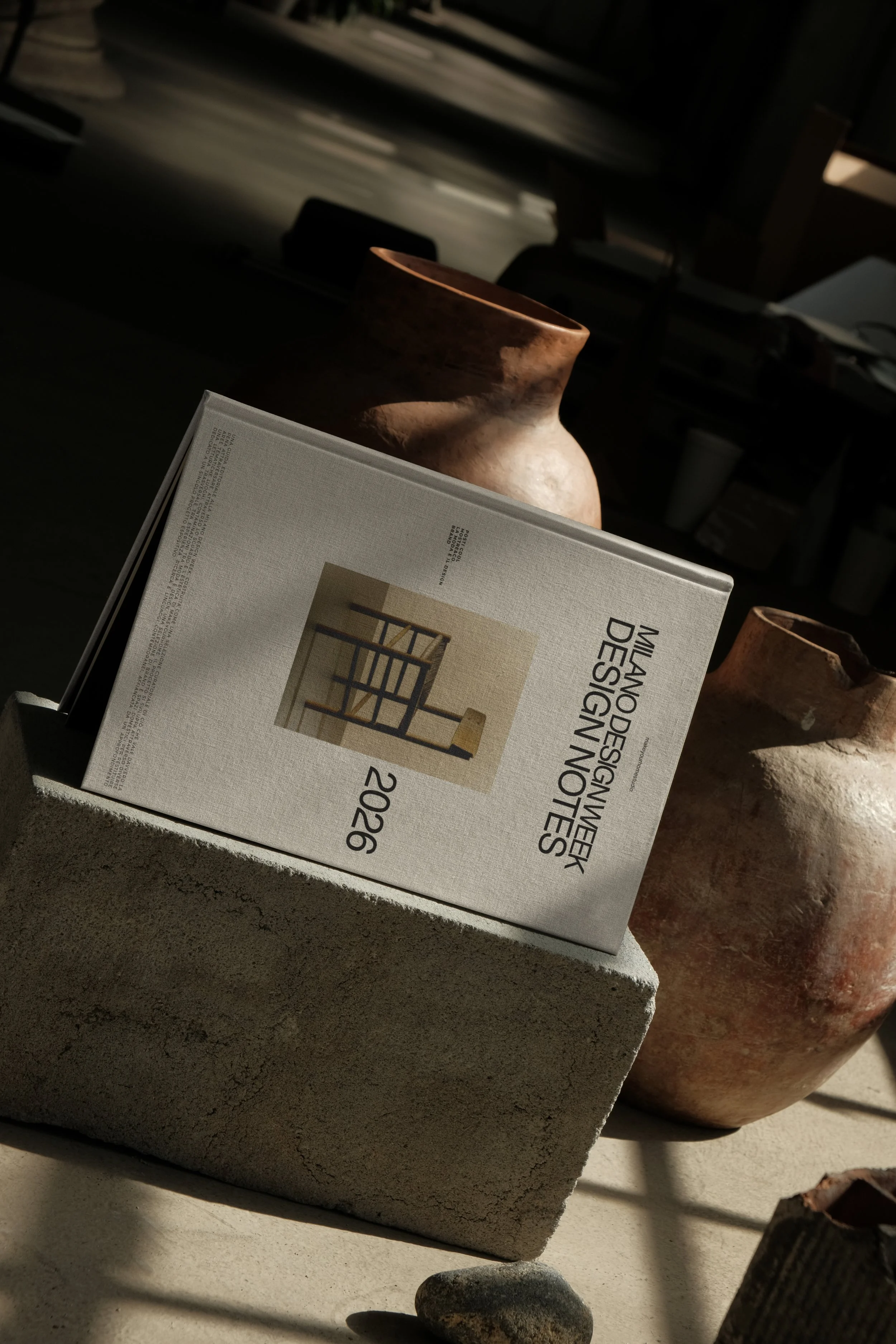 Libro sulla progettazione interiore appoggiato su un blocco di pietra, sfondo con scatole e oggetti vari in un ambiente con luce naturale.