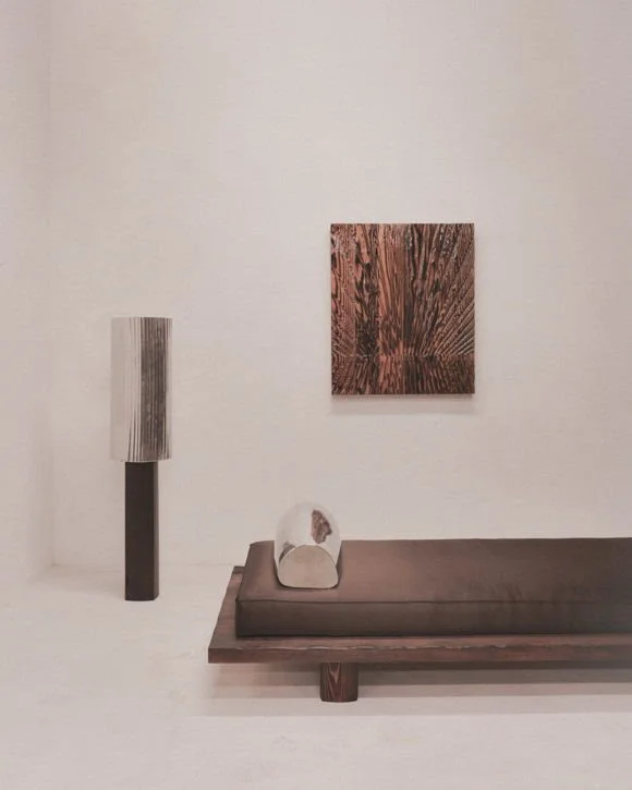 Interno di una stanza minimalista con una sedia a panca imbottita marrone, una lampada di design bianca e nera e un quadro astratto su parete bianca.
