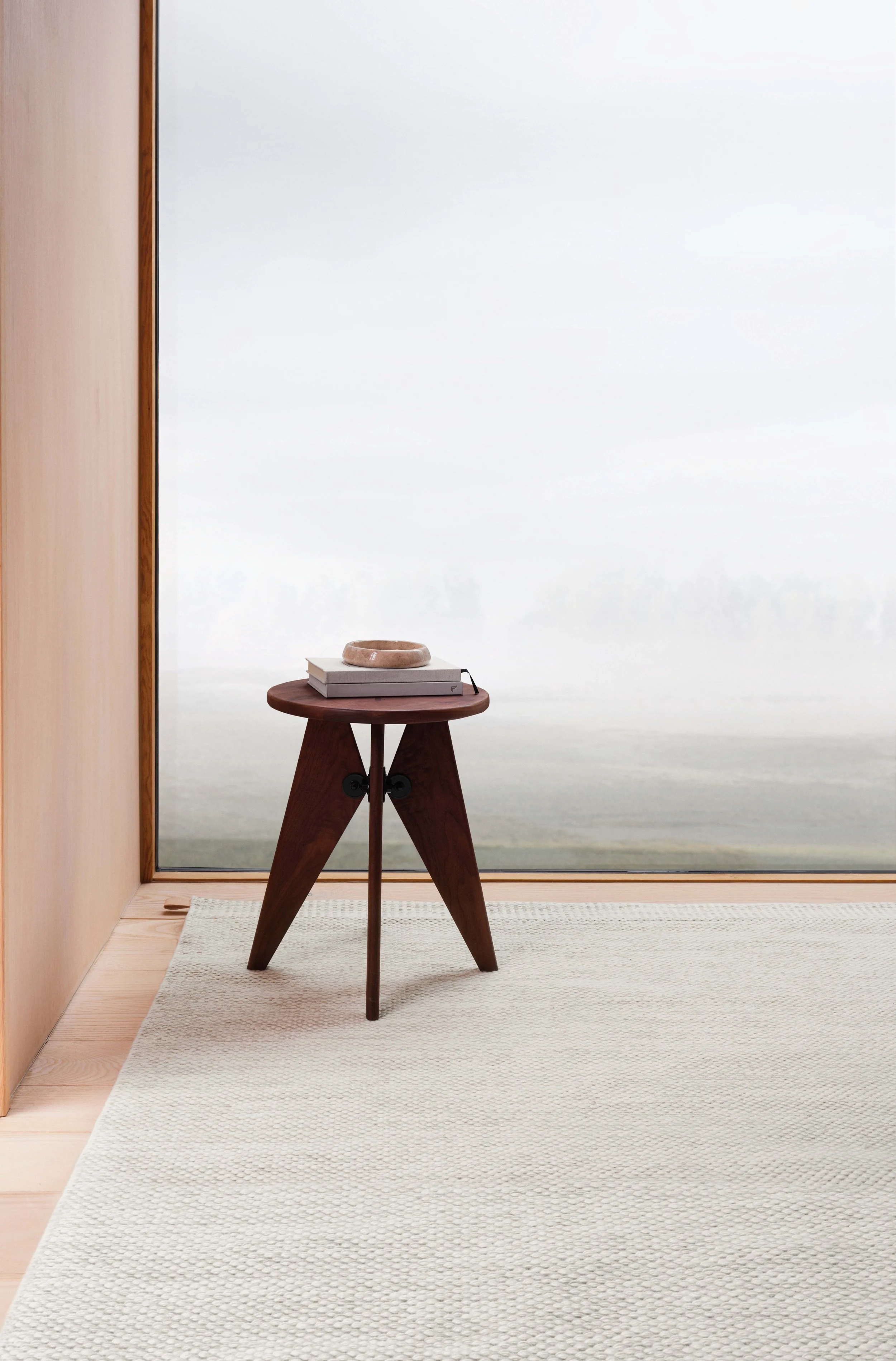 Un tavolo di legno scuro con tre gambe, con sopra due libri impilati e un recipiente o vaso di ceramica. L'ambiente è luminoso con pareti chiare, vicino a una grande finestra senza tende che lascia passare molta luce.