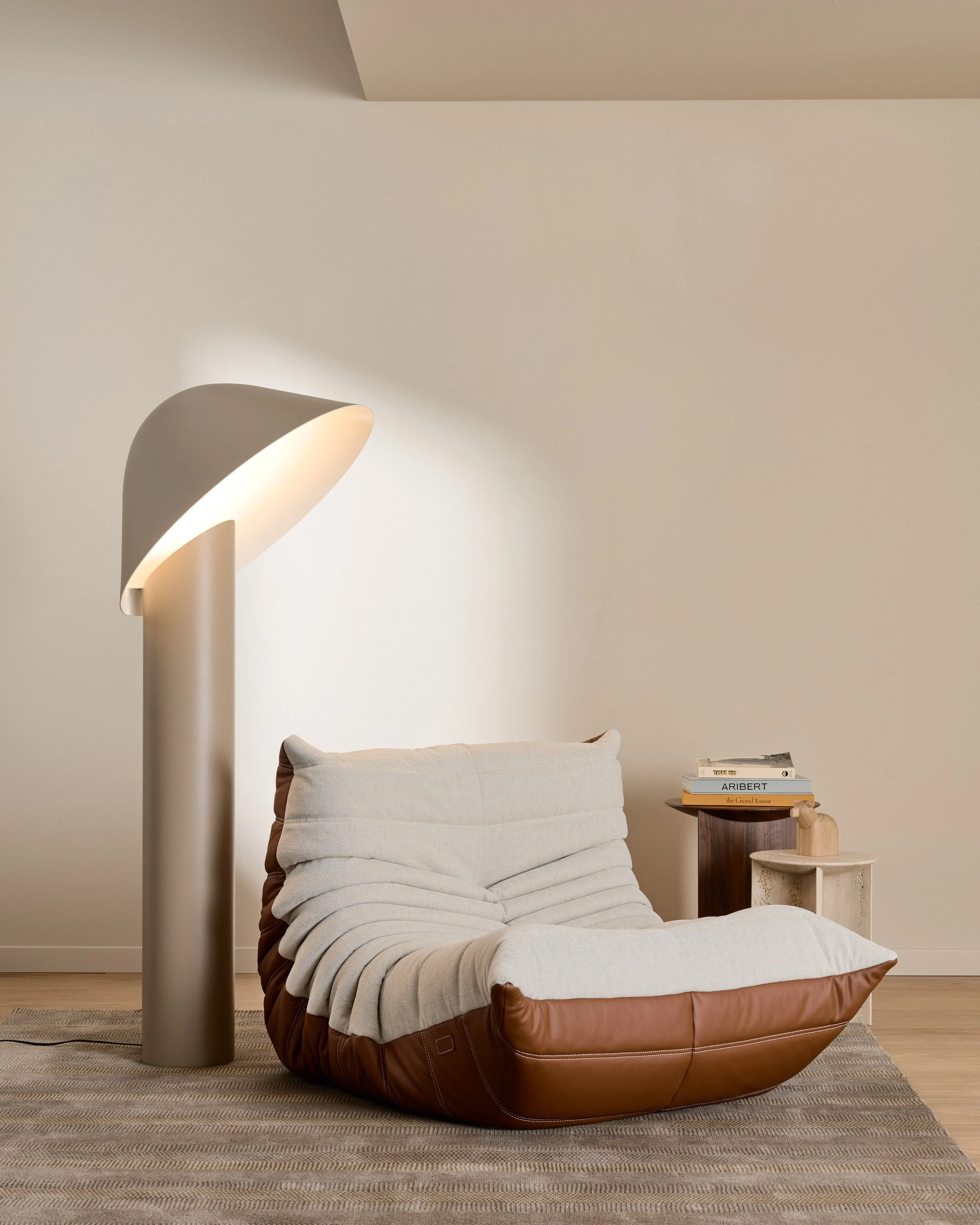 L'immagine mostra un angolo di una camera con un letto in pelle marrone e una coperta beige con cuscini. Accanto al letto ci sono alcune riviste impilate su un piccolo tavolino di legno, e sopra di esso si trova un complemento decorativo in legno con una piccola figura stilizzata. Sul muro alle spalle, un'illuminazione moderna e minimalista si estende lungo l'angolo, creando un'atmosfera calda e accogliente.