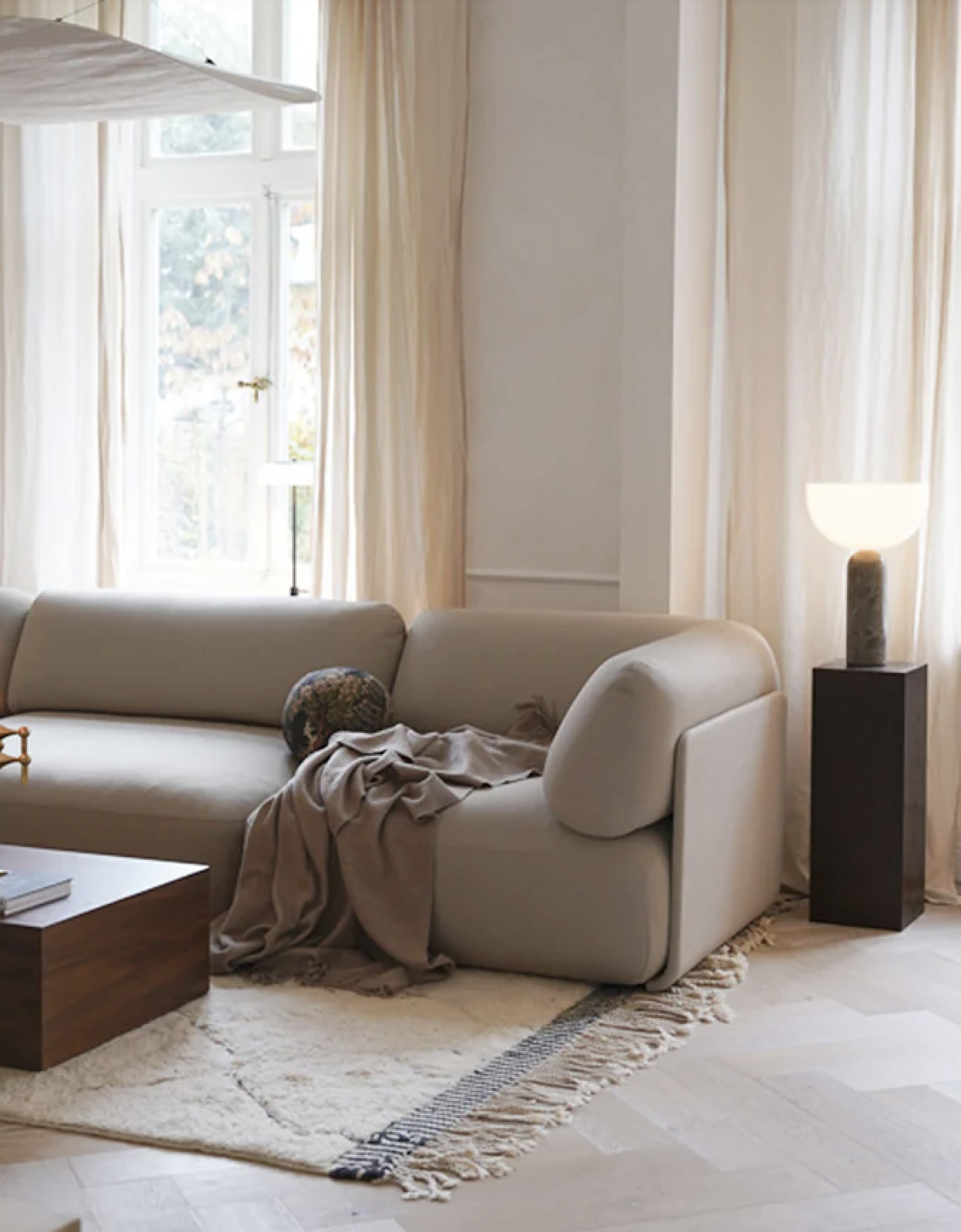 Soggiorno con divano beige, tappeto chiaro, finestra con tende colori chiari, lampada da terra con base nera e paralume bianco, tavolino di legno con libri.