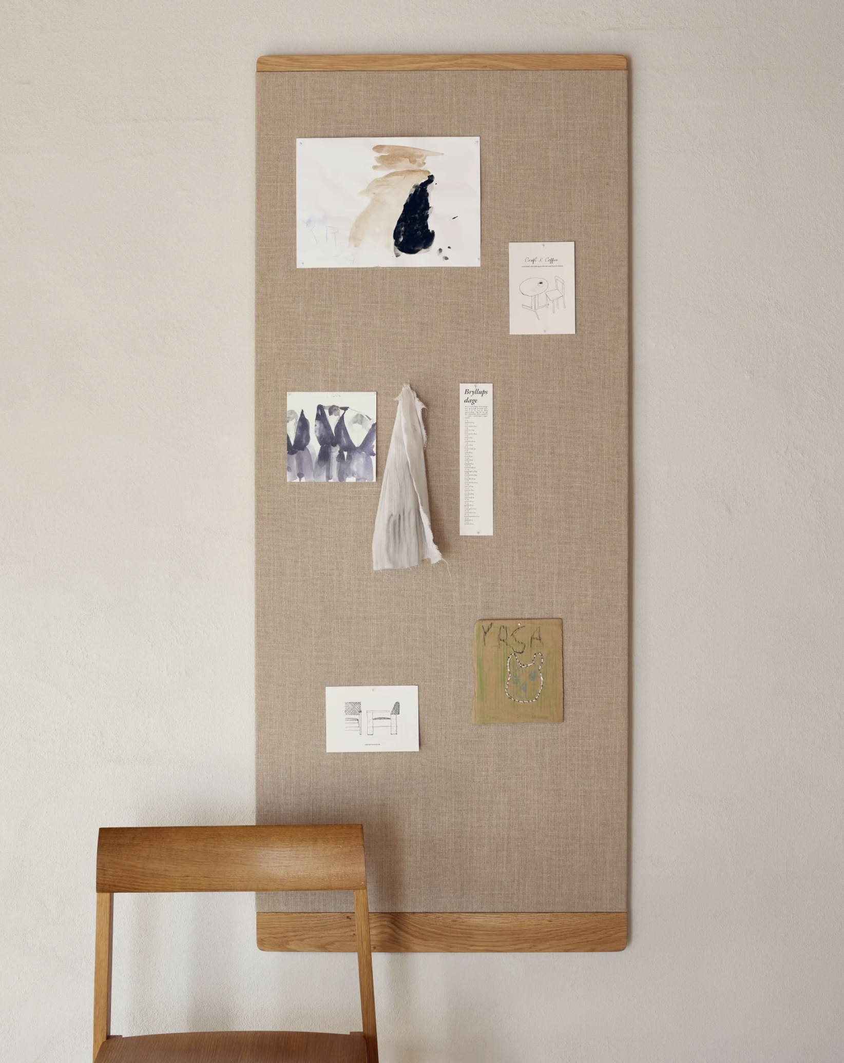 Bacheca con vari appunti, disegni e fotografie, appesa al muro, con una sedia di legno davanti.