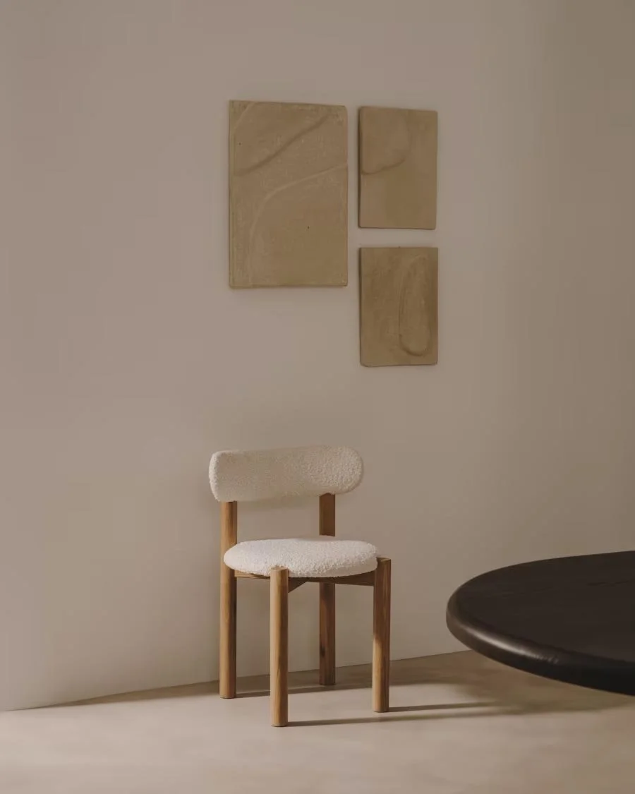 Sedia con struttura in legno e rivestimento in tessuto bianco, posizionata accanto a un tavolo rotondo nero, in un ambiente minimalista con parete decorata con tre quadri di arte astratta in toni beige.