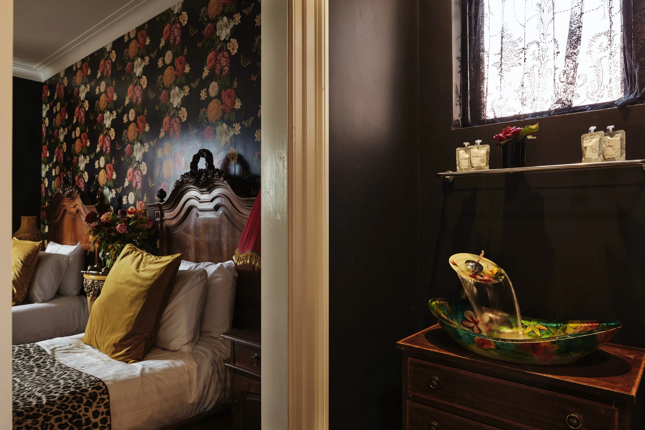Interior of a vintage-style bedroom with floral wallpaper, wooden headboards, yellow pillows, and a leopard print blanket. Adjacent bathroom features colorful glass basin sink and toiletries.