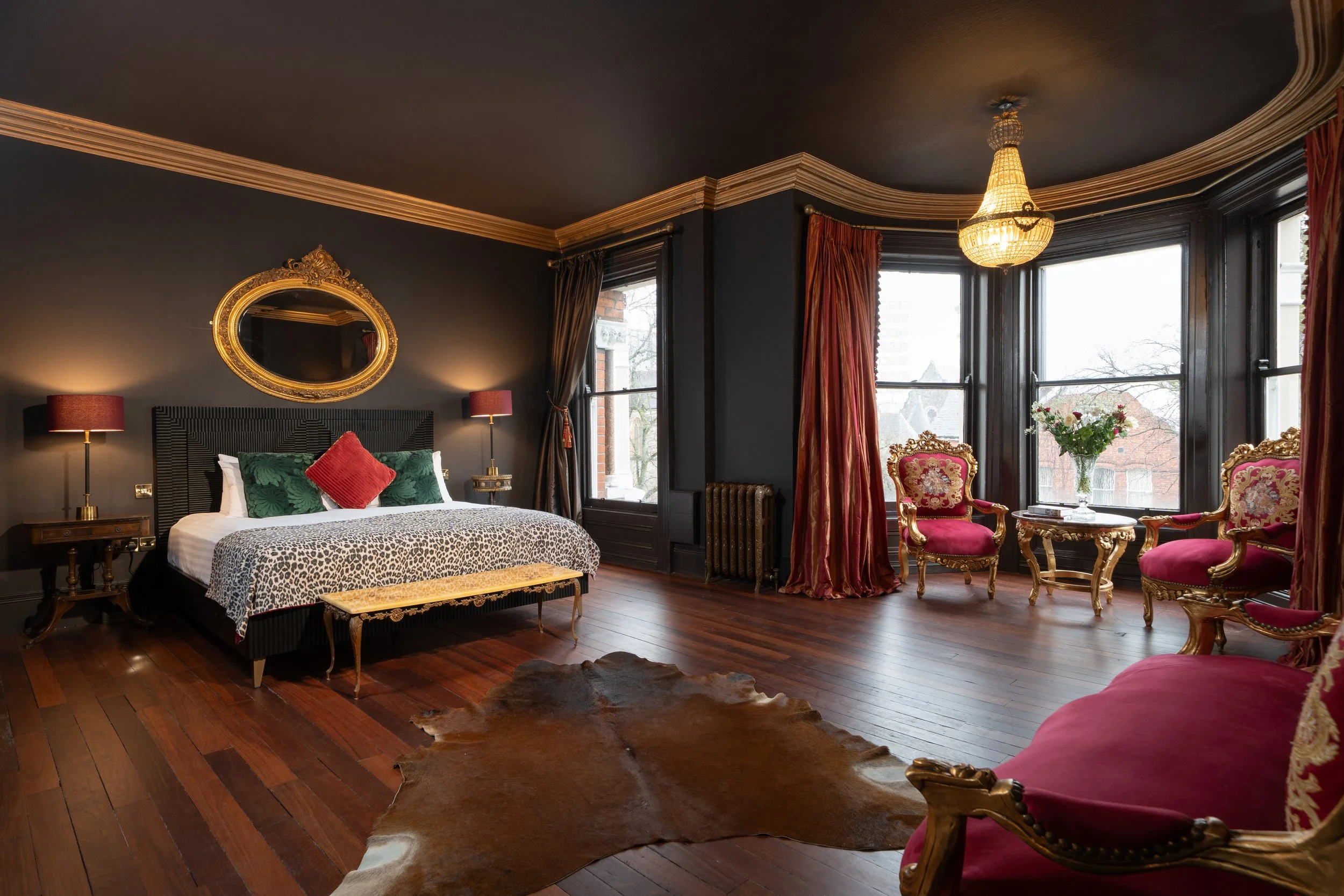 Luxurious bedroom with a king-size bed, ornate mirror, elegant chandelier, red chairs, and a cowhide rug.
