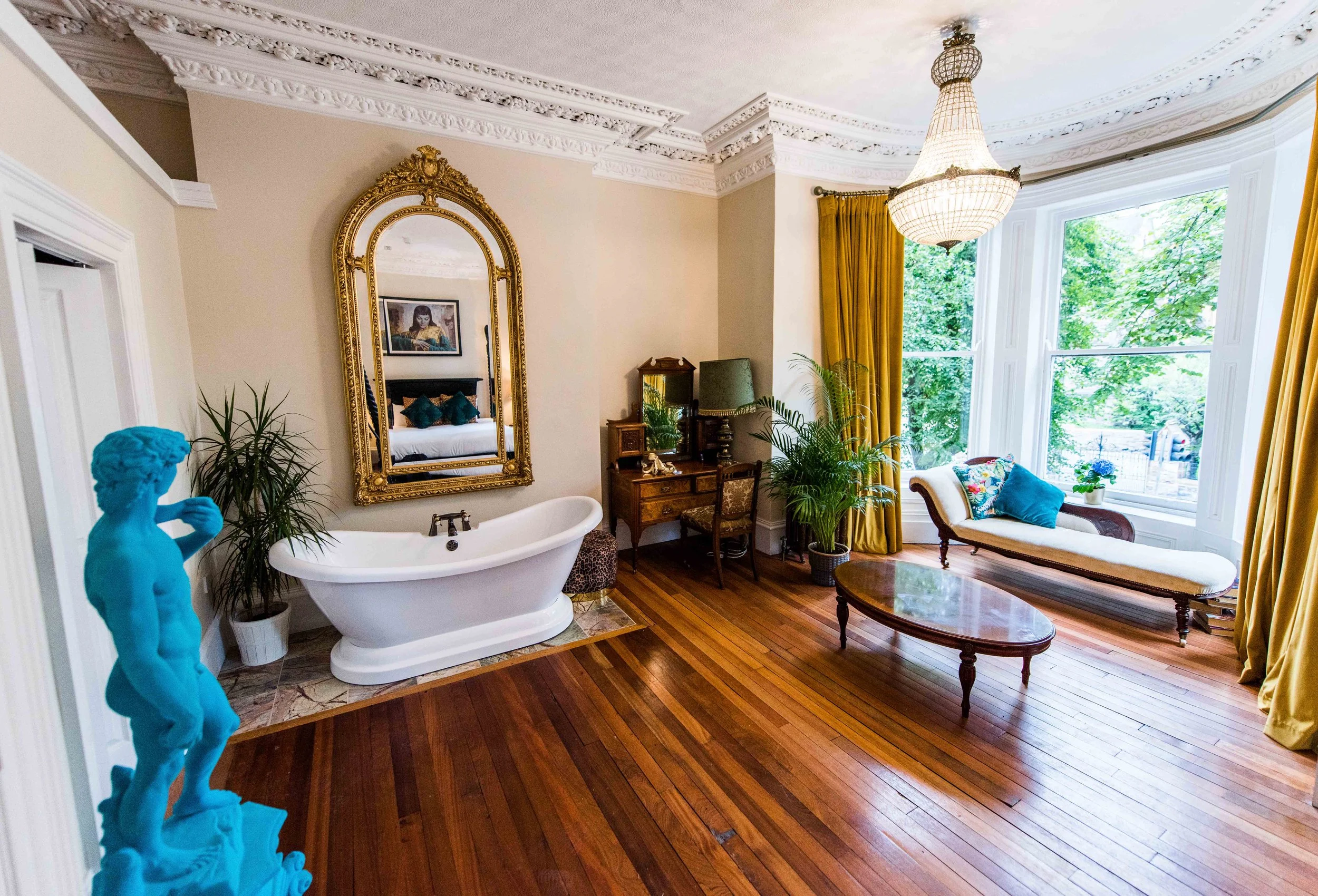 Elegant bathroom with a freestanding tub, gold ornate mirror, chandelier, hardwood floors, and large window with yellow curtains, featuring a chaise lounge and plants.