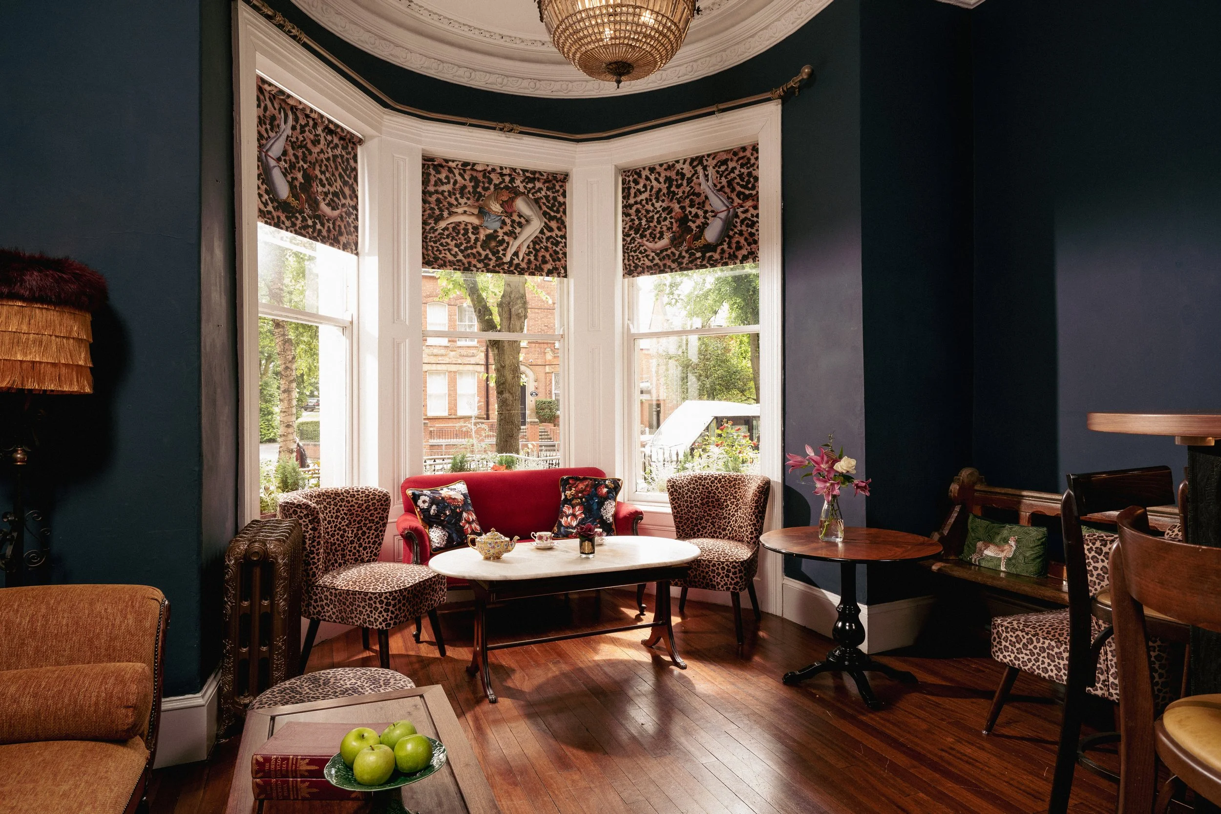 A cozy living room with dark blue walls and a large bay window. The room features a red sofa, leopard print chairs, and a wooden coffee table with green apples. A vintage-style chandelier hangs from the ceiling, and there are floral accents on pillow