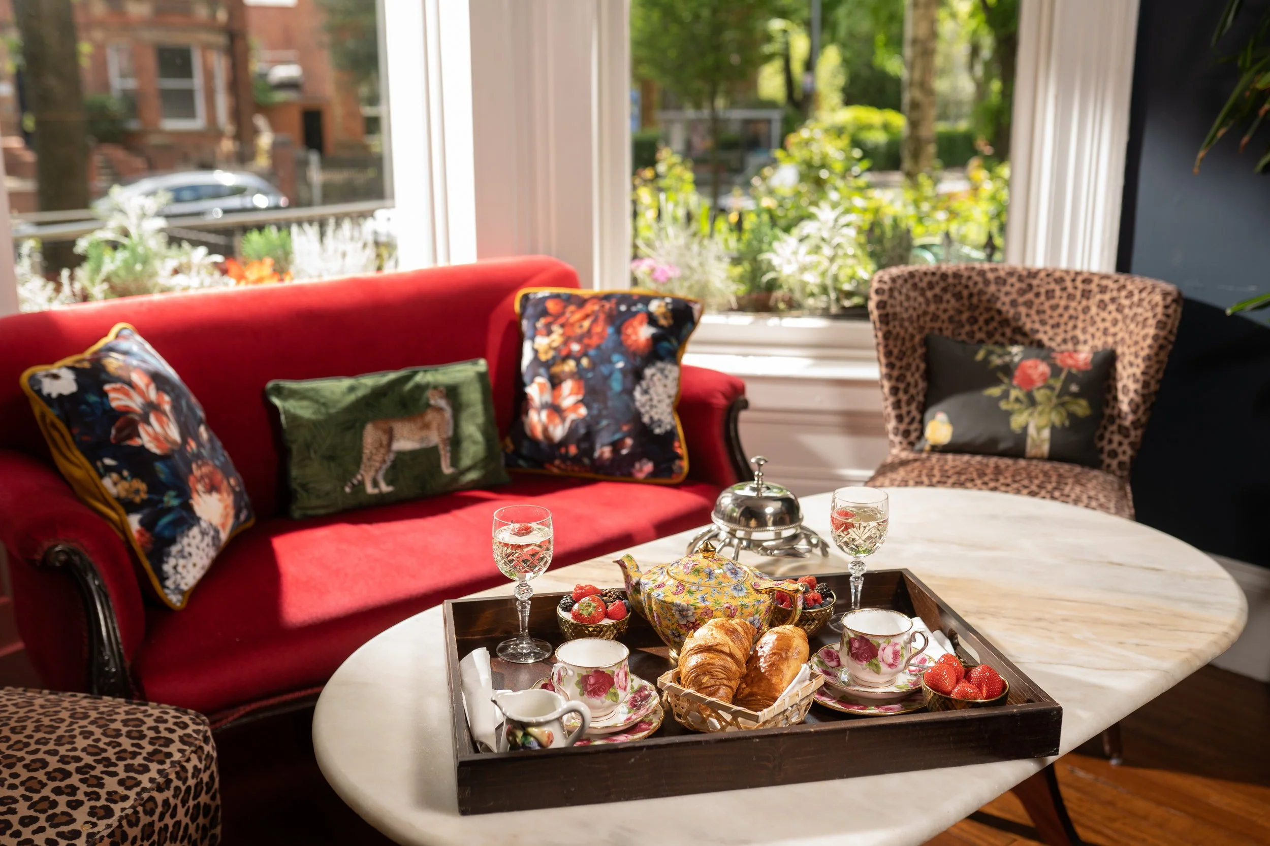 Cozy living room setup with red sofa and leopard-print chair, floral and cheetah pillows, a marble table with a breakfast tray featuring tea set, croissants, and strawberries.