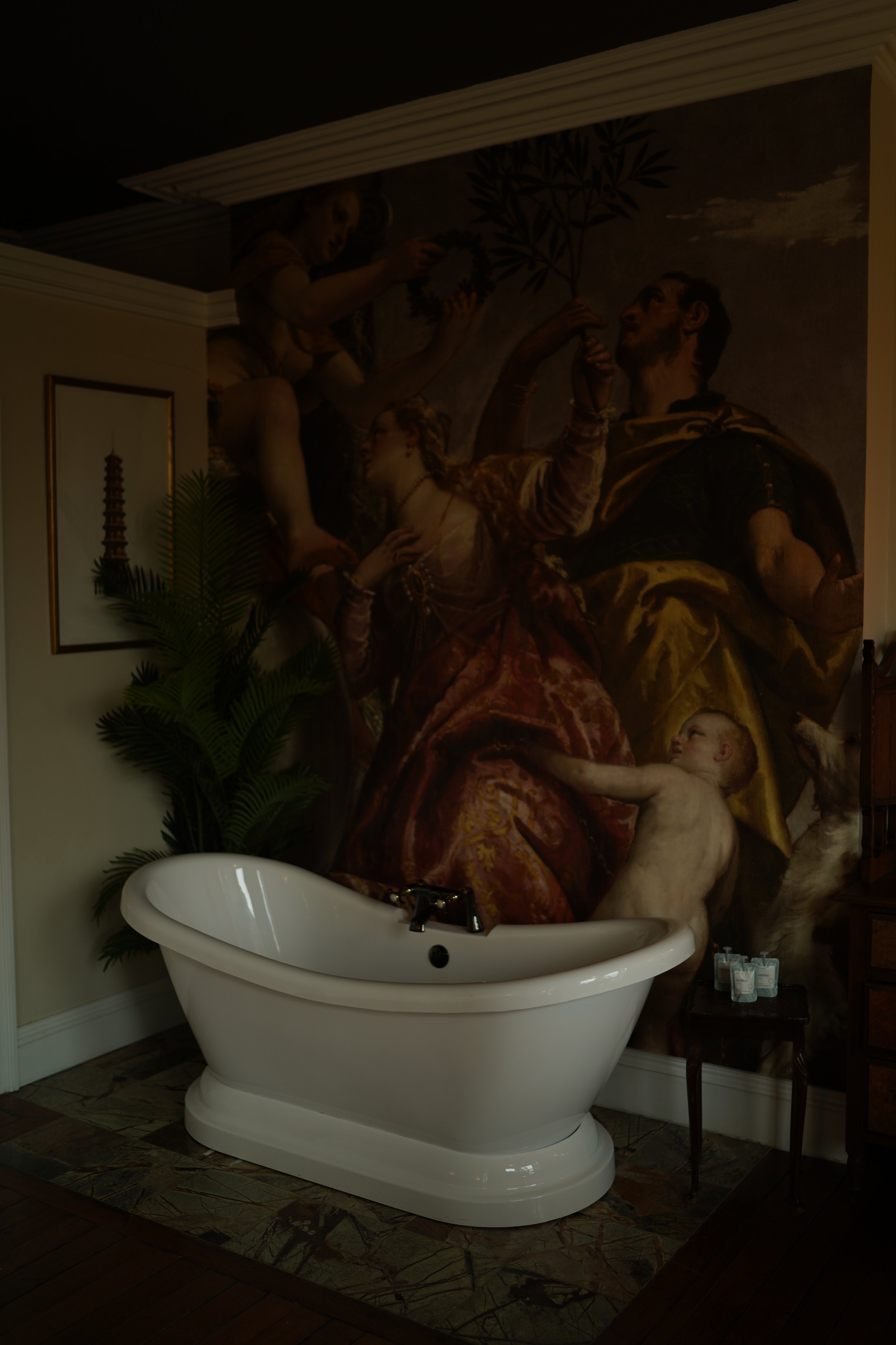 Elegant bathroom with a white freestanding bathtub, classical wall mural depicting historical figures, and a potted plant beside the tub.