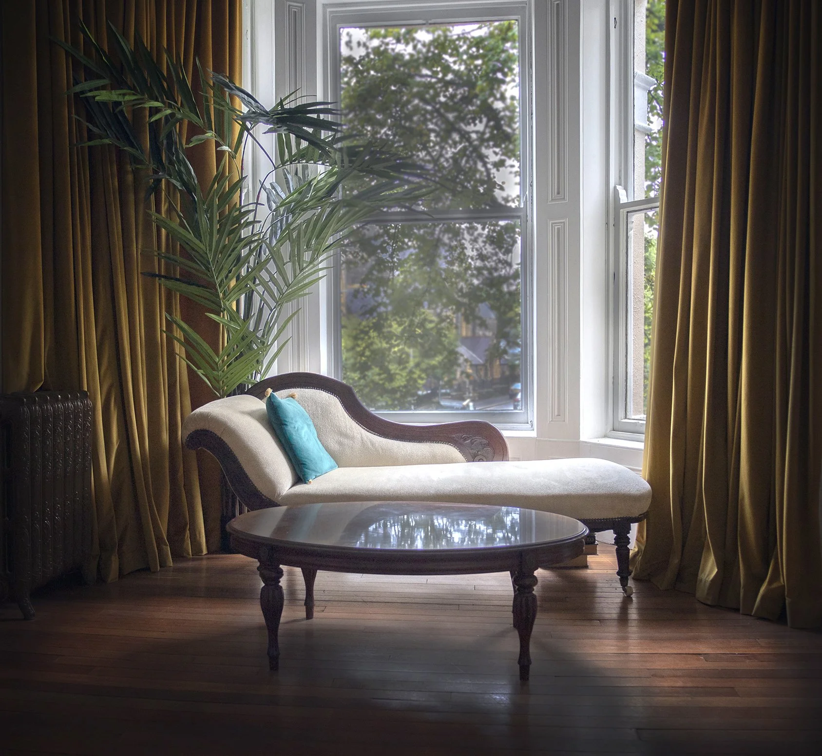 Elegant living room with a chaise lounge, circular coffee table, large windows, and a tall plant, featuring golden curtains and wooden flooring.