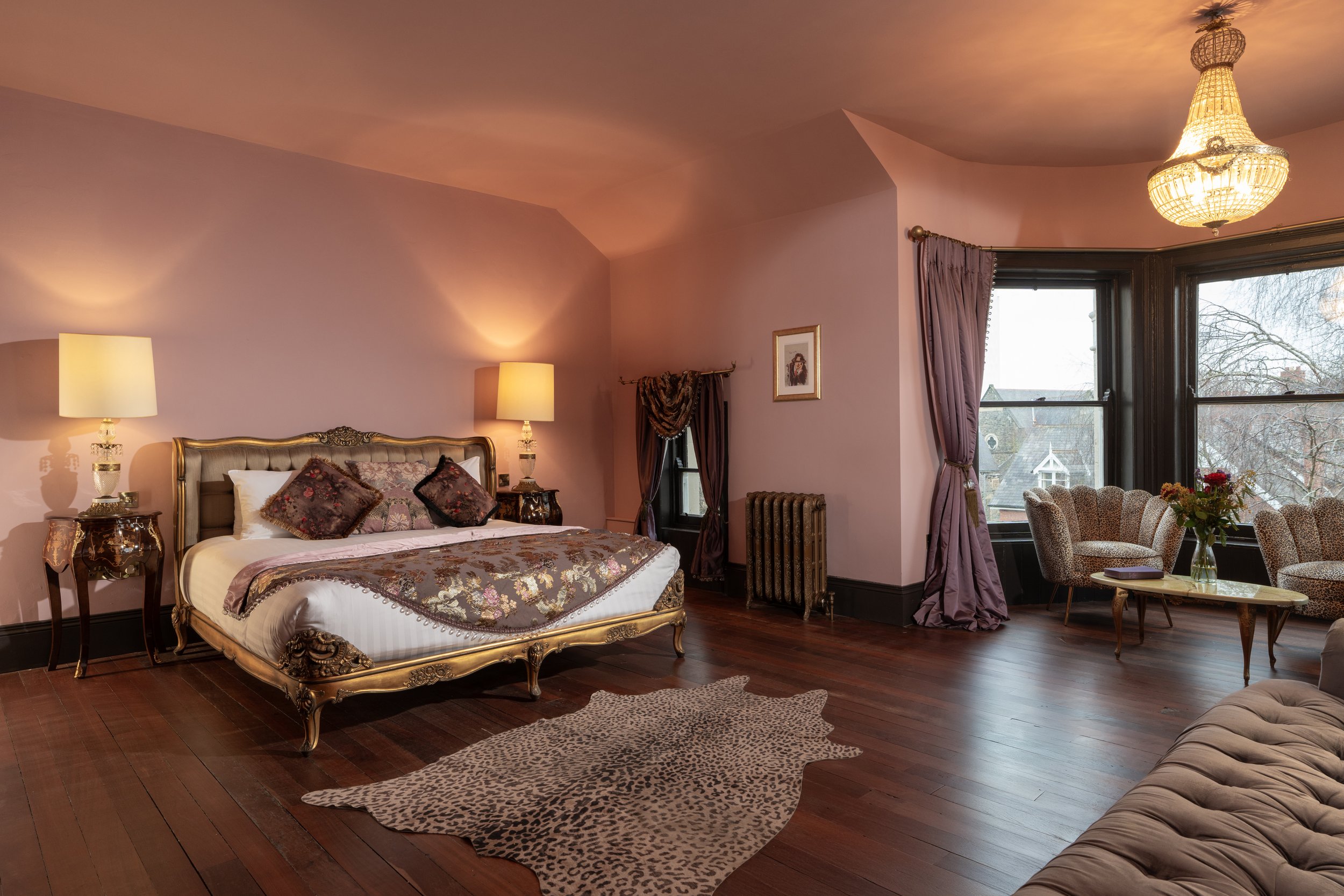Luxurious bedroom with ornate bed, floral bedding, bedside tables with lamps, a plush area rug, vintage-style chairs by a window, and a chandelier.