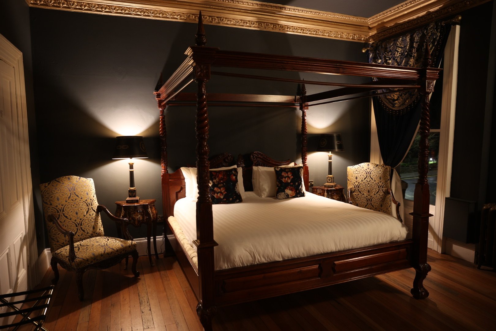 A luxurious bedroom with a wooden four-poster bed, patterned chairs, bedside tables, and decorative pillows, illuminated by lamps.