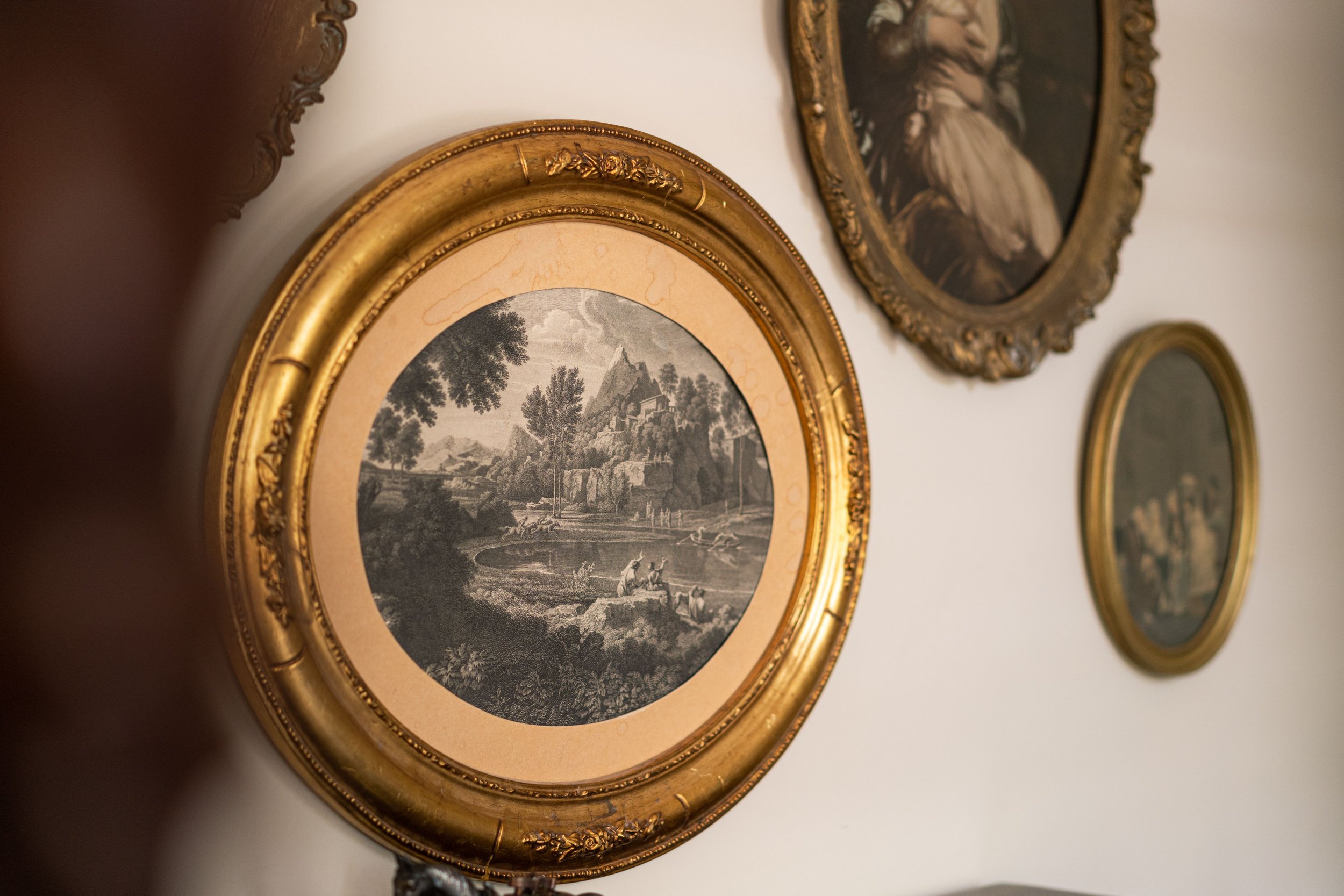 Framed oval paintings on a wall, featuring a classical landscape in the foreground.