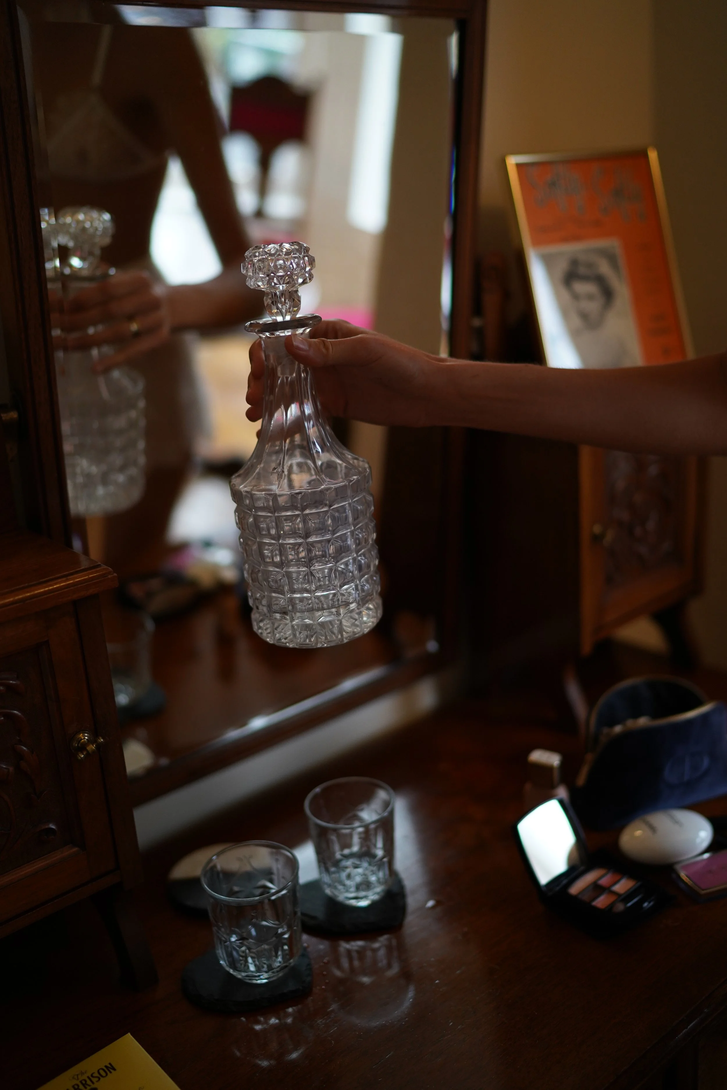 Person holding a crystal decanter in front of a mirror, with two glasses on coasters nearby on a wooden vanity.