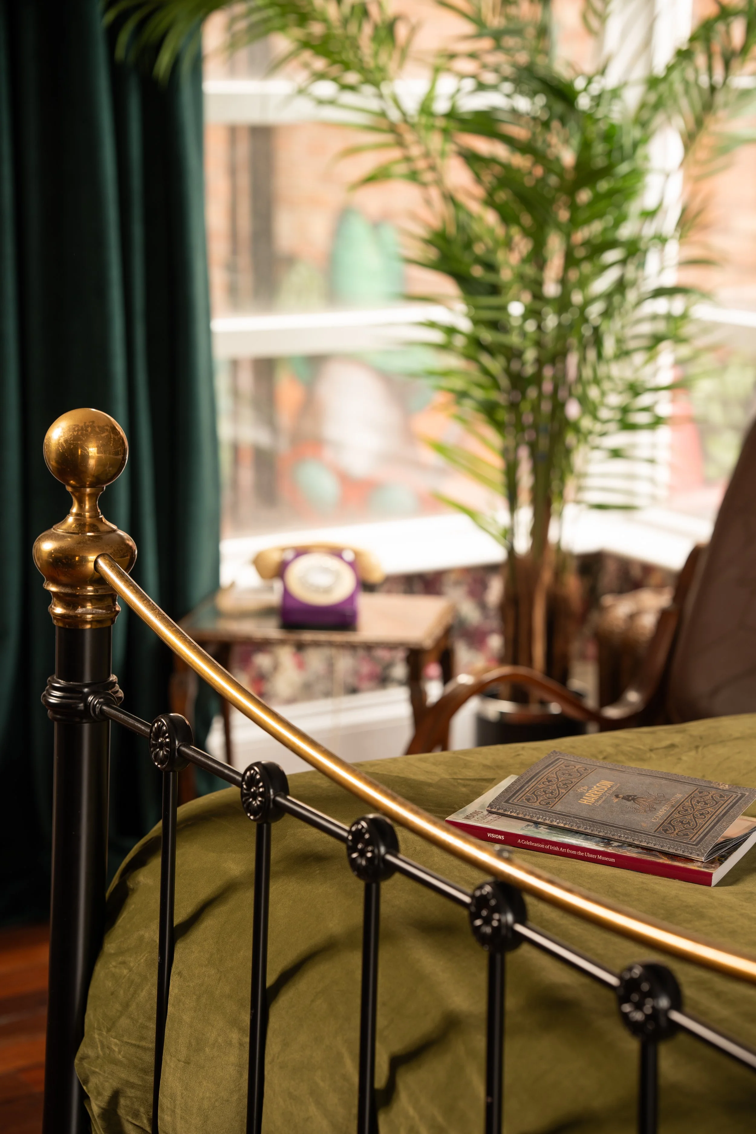 A cozy bedroom with a metal bed frame, green bedspread, and books on top. In the background, a side table with a vintage rotary telephone, a large potted plant, a window with sunlight, and a chair with a patterned cushion are visible.