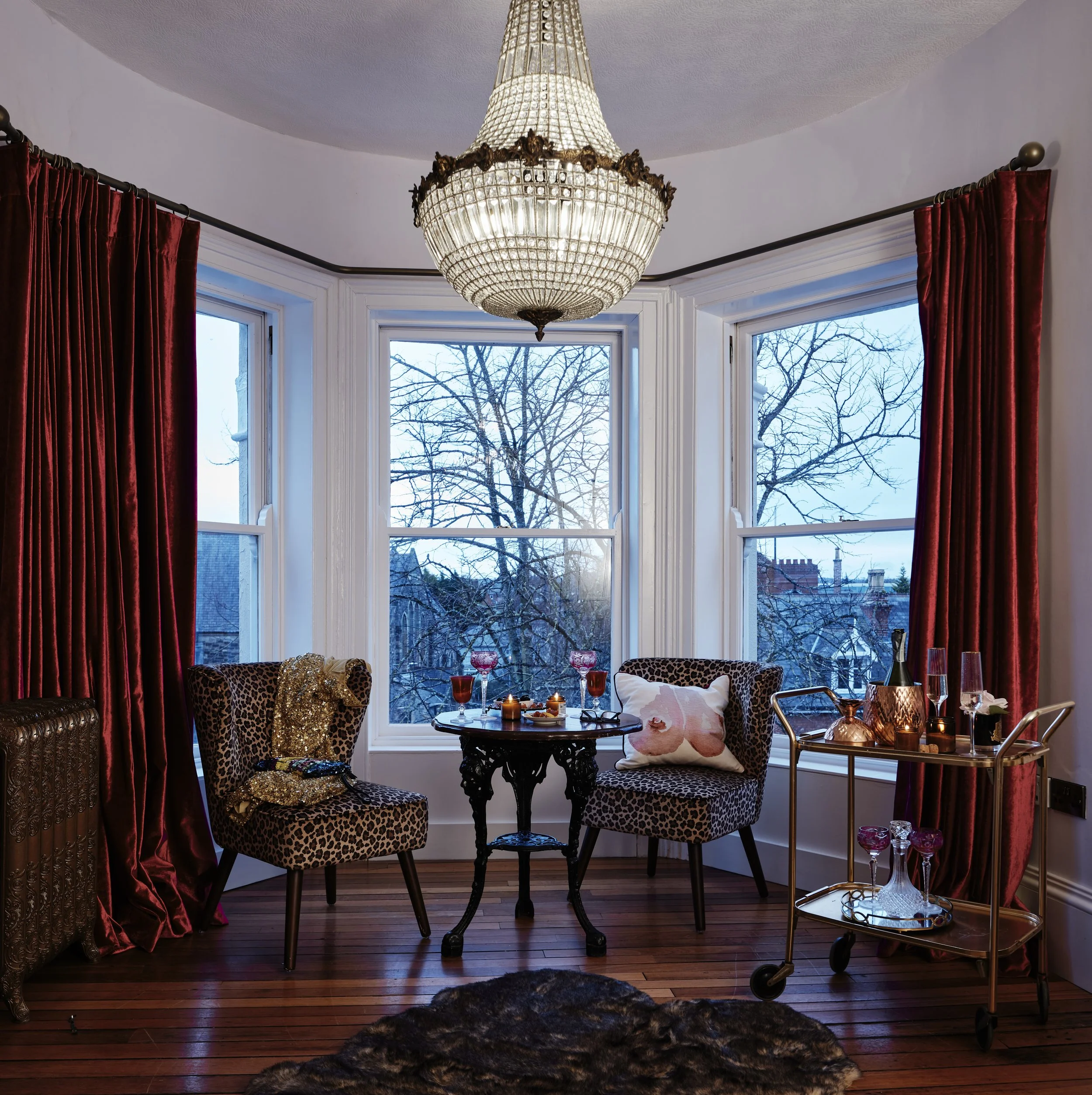 A cozy room with a vintage chandelier, two leopard print chairs, a small round table with drinks, red curtains, and a drink cart. The large windows show a tree and rooftops outside.