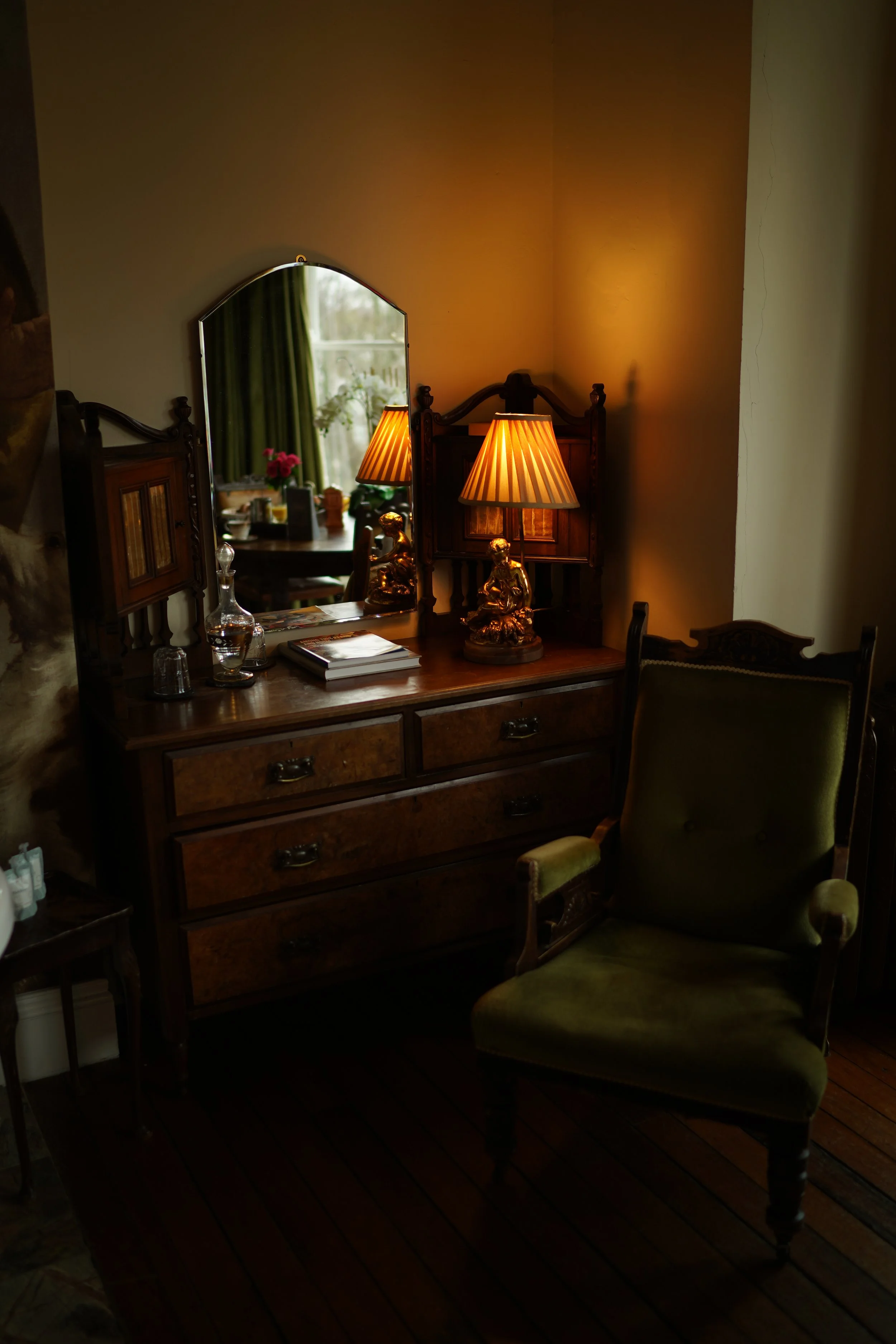 Vintage-style room with a wooden dresser, mirror, table lamp, and green upholstered chair.