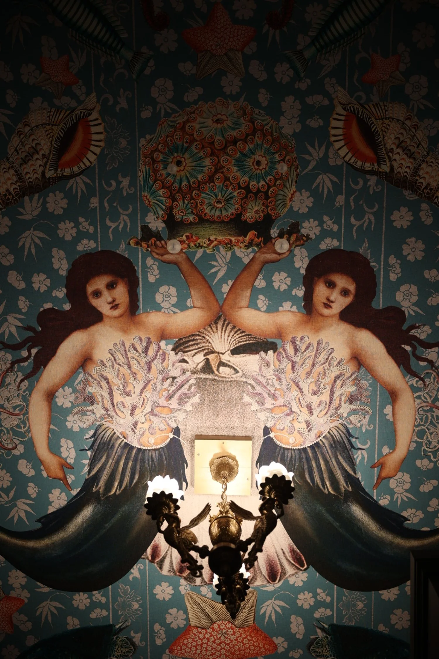 Artistic wallpaper featuring two mirrored mermaids with shells and sea motifs, showing a vintage chandelier in the foreground.