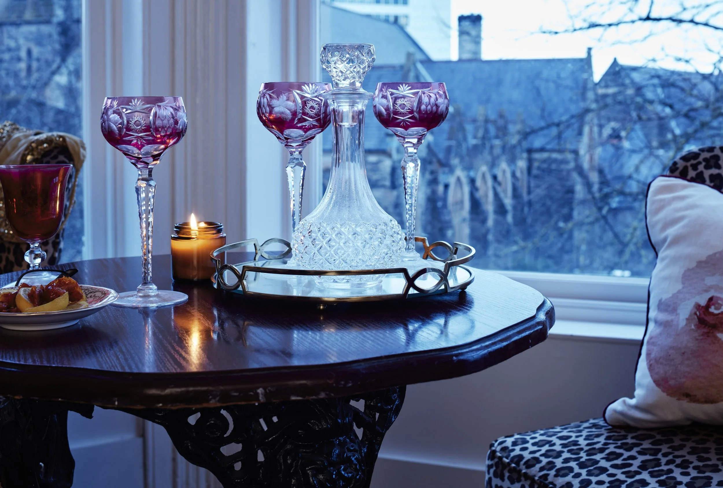 Elegant dining setup with crystal decanter, red wine glasses, lit candle, and plate of fruit on wooden table by a window.