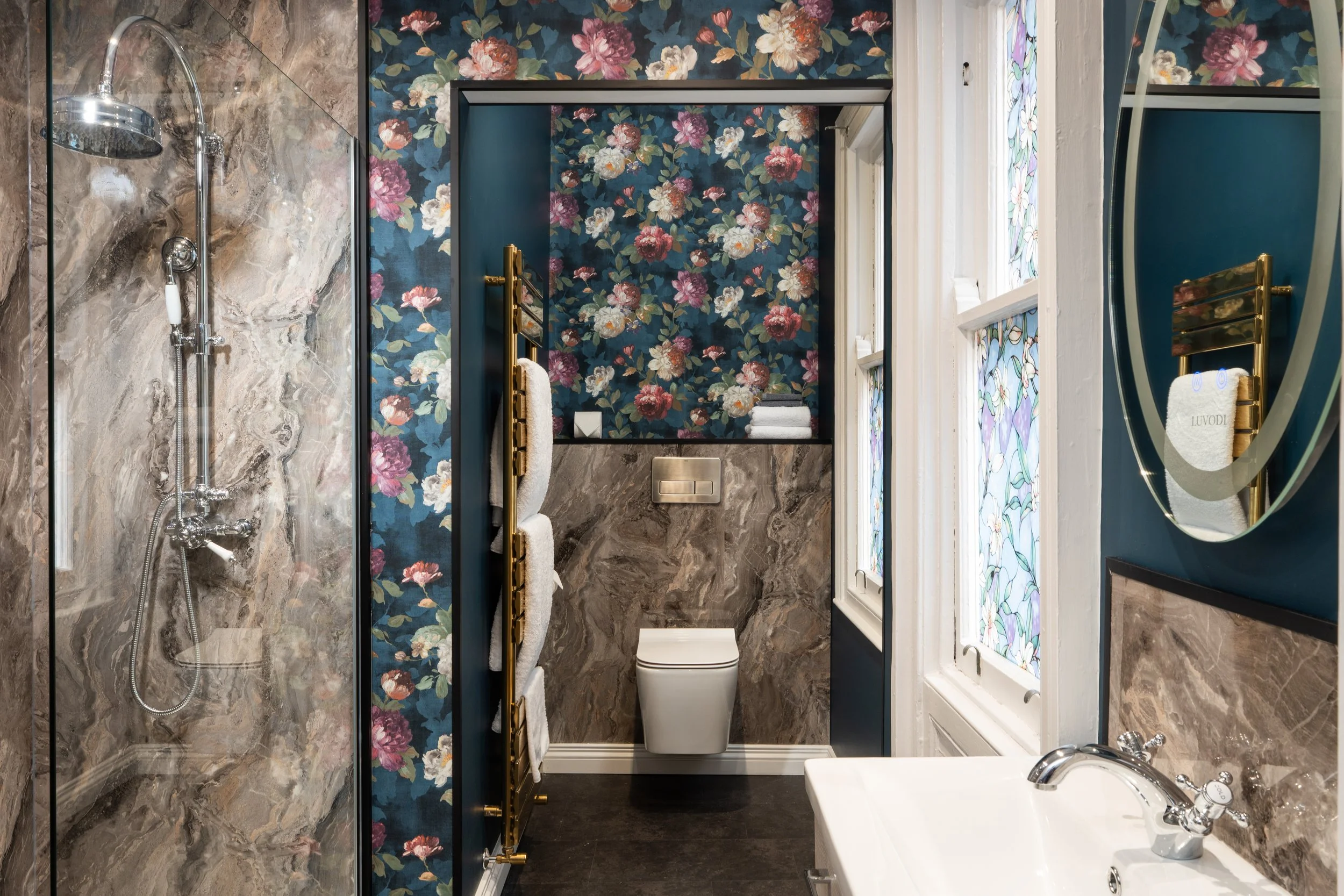 Elegant bathroom with floral wallpaper, marble-style tiles, a modern shower, glass screen, wall-mounted toilet, gold towel rack with white towels, stained glass windows, and a sleek sink with mirror.
