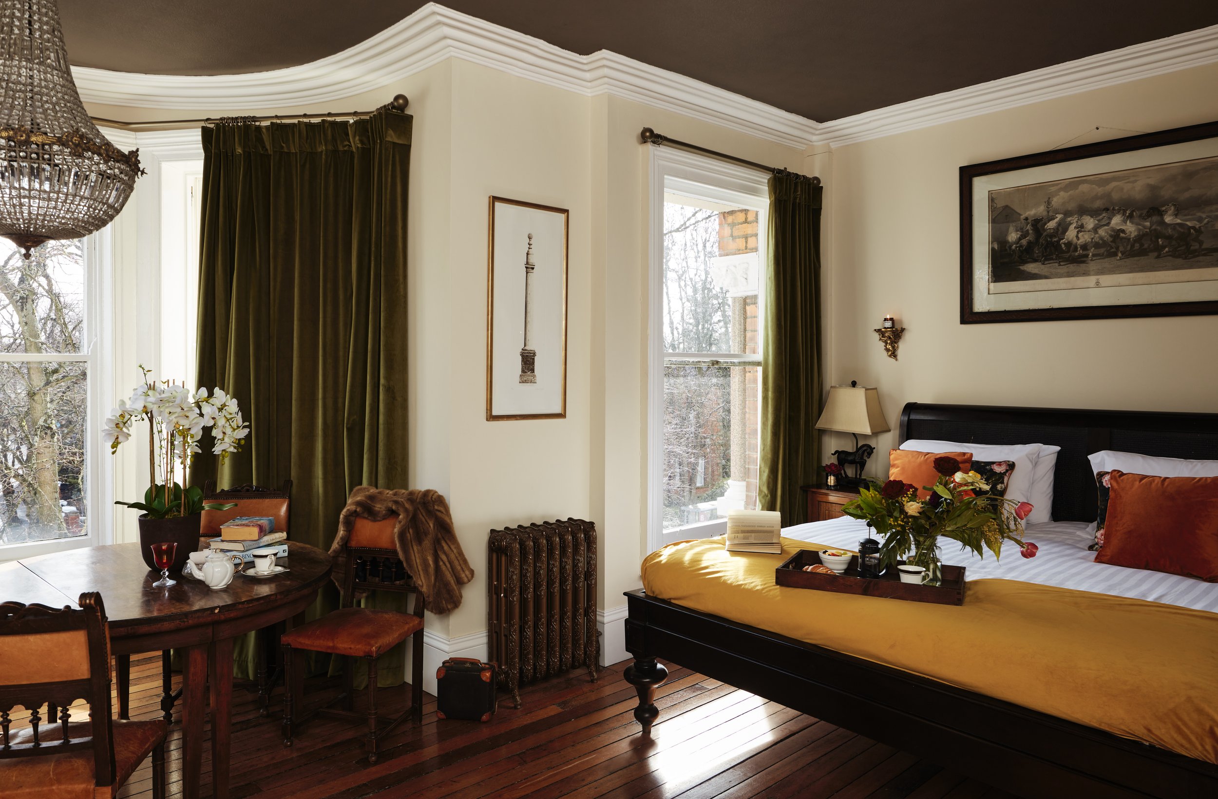 Elegant bedroom interior with a vintage chandelier, wooden furniture, and a bed with yellow bedding. Features include green curtains, a framed painting, decorative pillows, and fresh flowers on a tray.