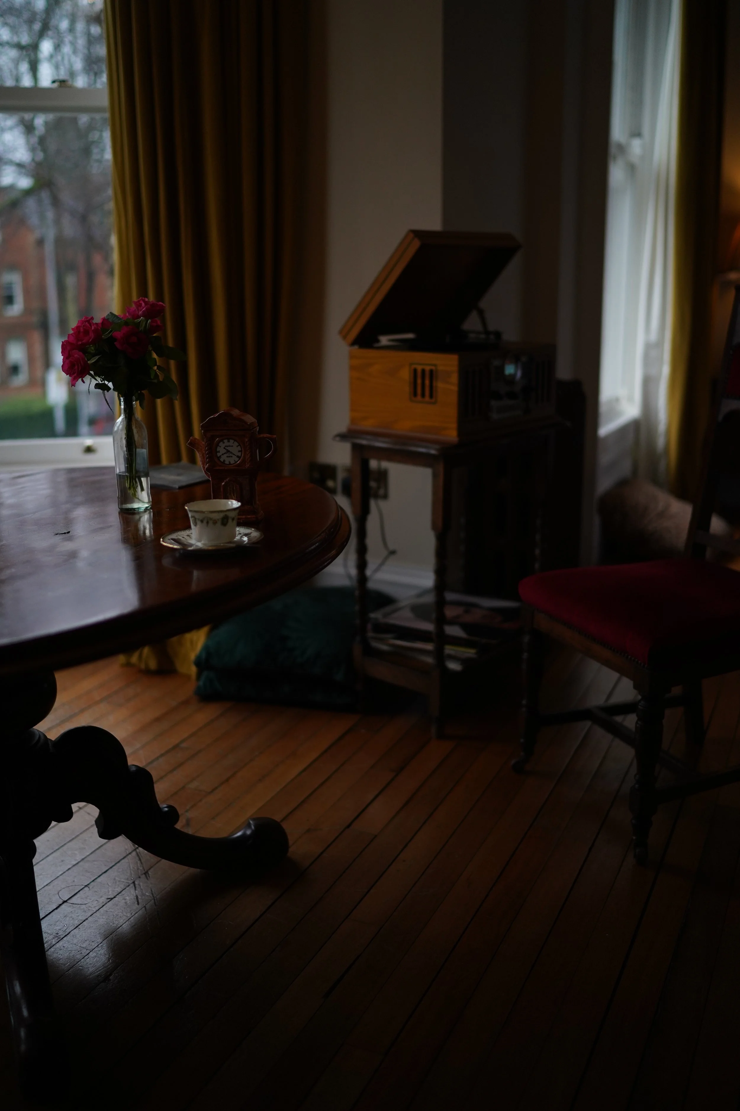 Room interior with vintage phonograph, wooden table, flowers in a vase, antique clock, and teacup. Yellow curtains frame the window, creating a warm ambiance.