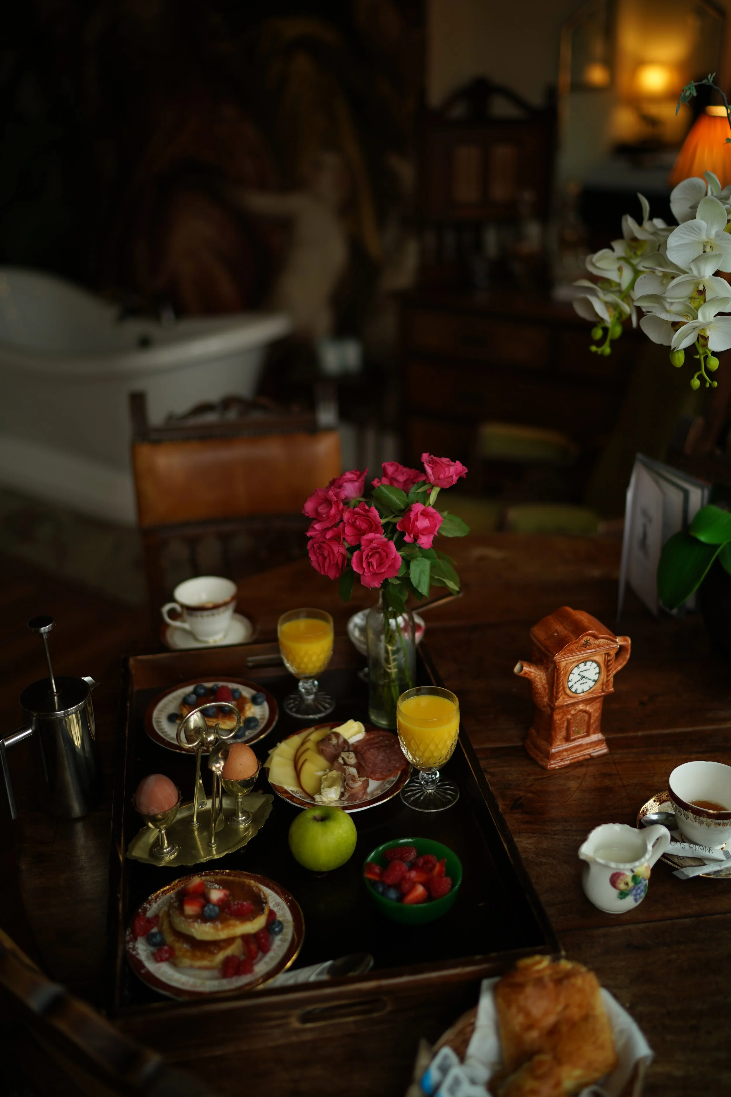 Breakfast tray with pancakes, eggs, fruit, orange juice, coffee, and flowers on a wooden table.