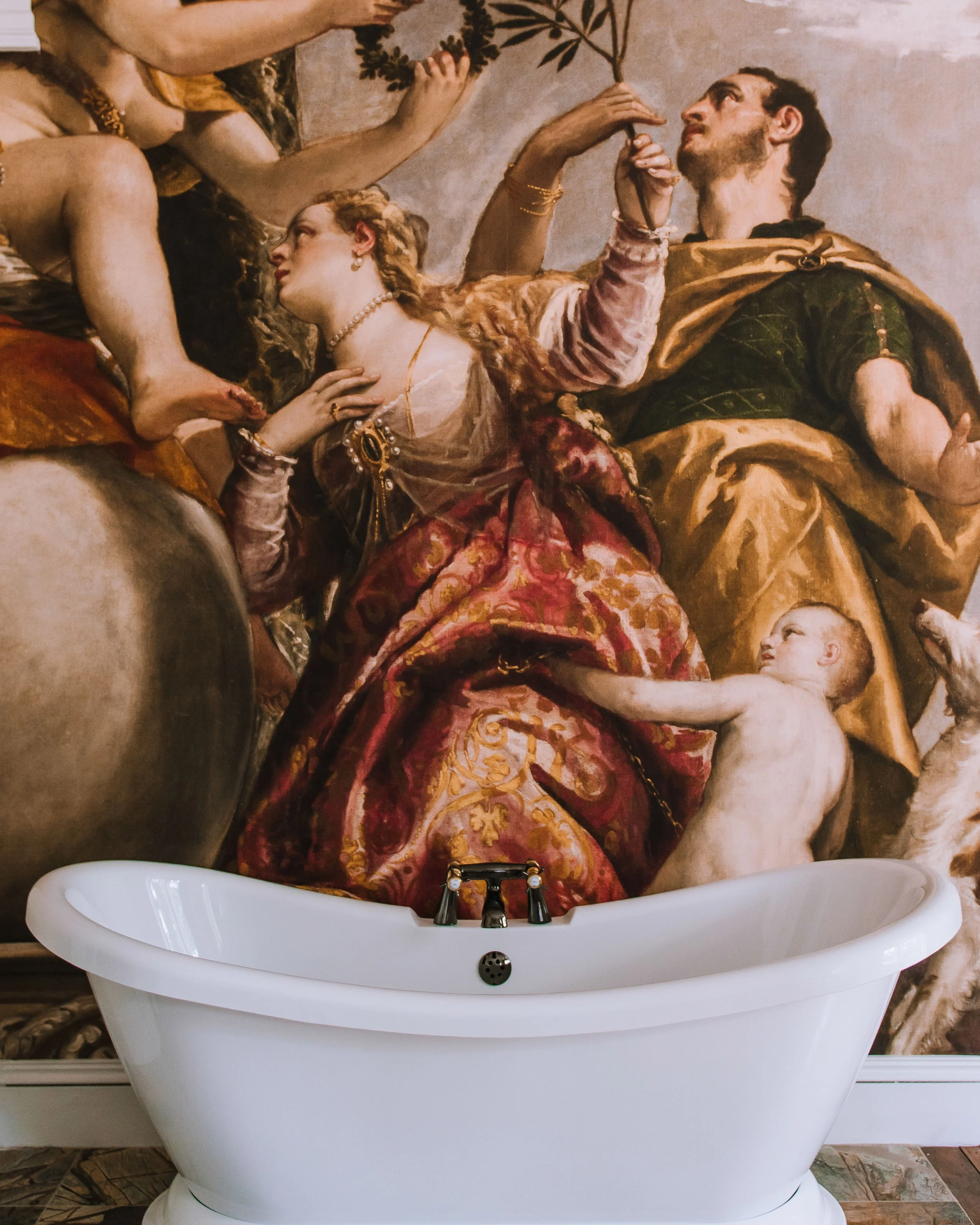 A white freestanding bathtub is placed in front of a classical Renaissance-style mural depicting figures in elaborate clothing and dramatic poses. The mural includes a woman in a red and gold patterned robe, a man in a green and gold outfit, and a ch