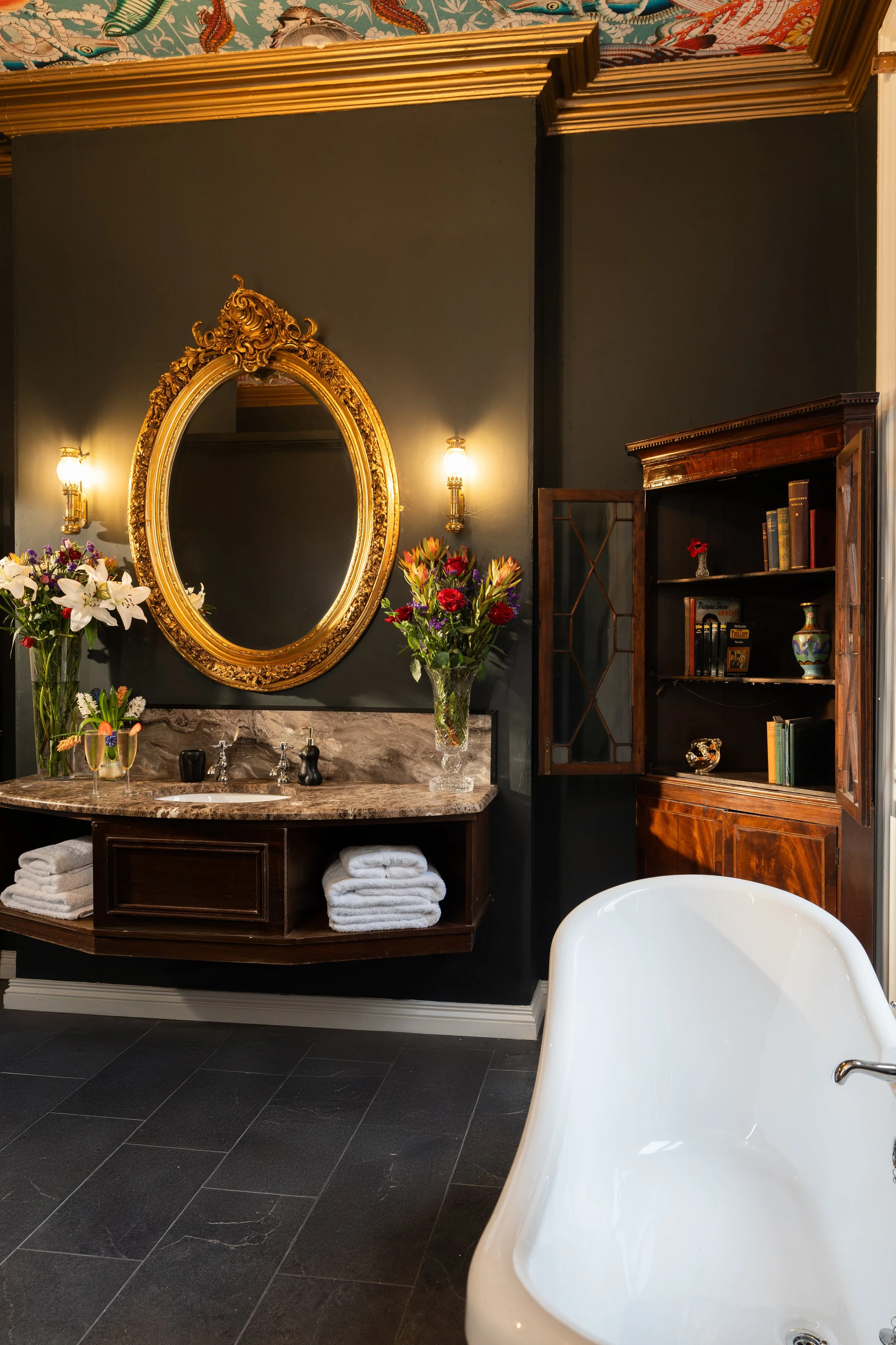 Luxurious bathroom with a vintage gold oval mirror, ornate lights, decorative dark walls, stone countertop, wooden shelving, fresh towels, floral arrangement, and a white bathtub.