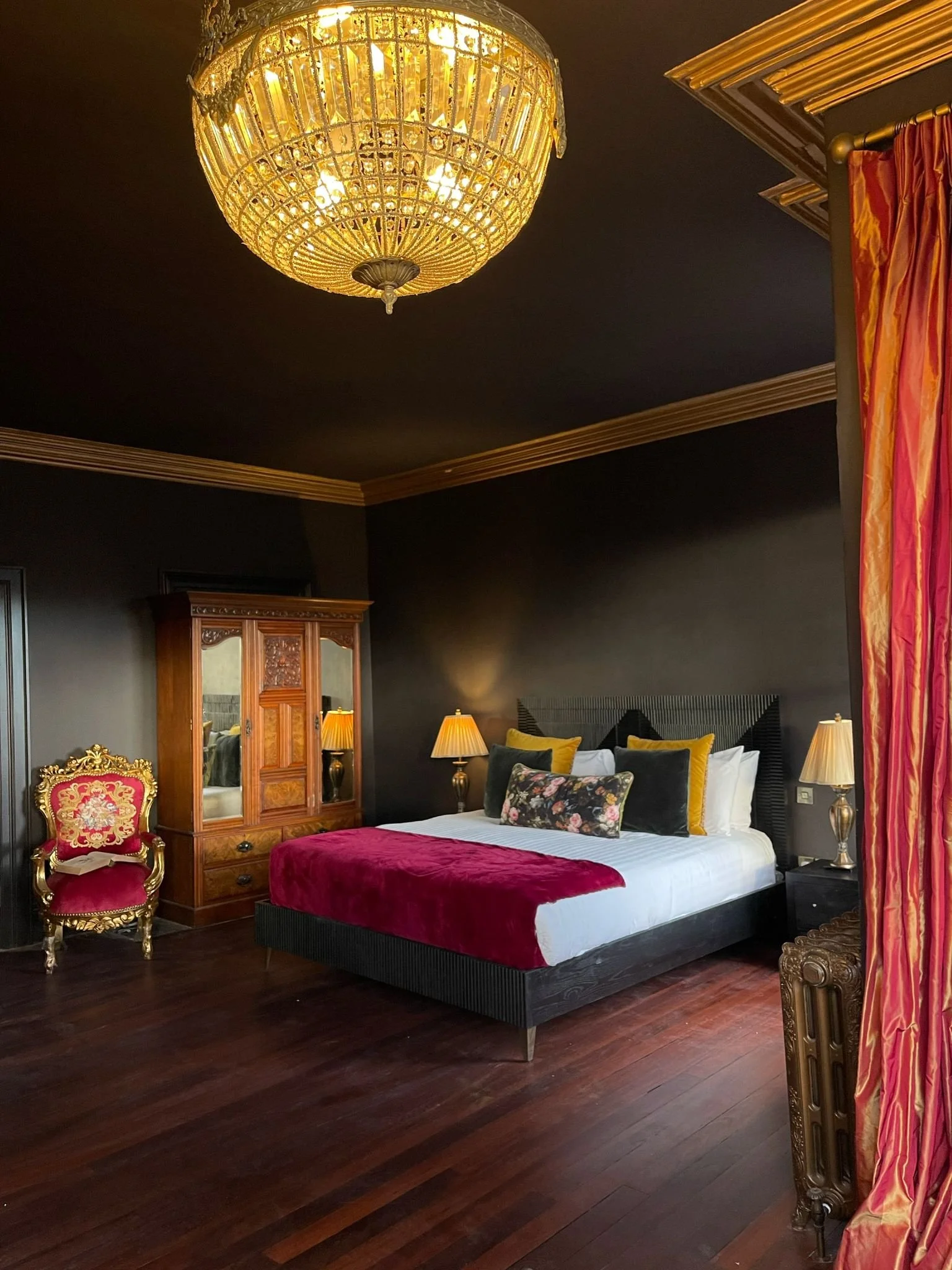 Luxurious bedroom with ornate chandelier, dark walls, and wooden furniture, featuring a large bed with red and gold accents, a vintage armchair, and red curtains.
