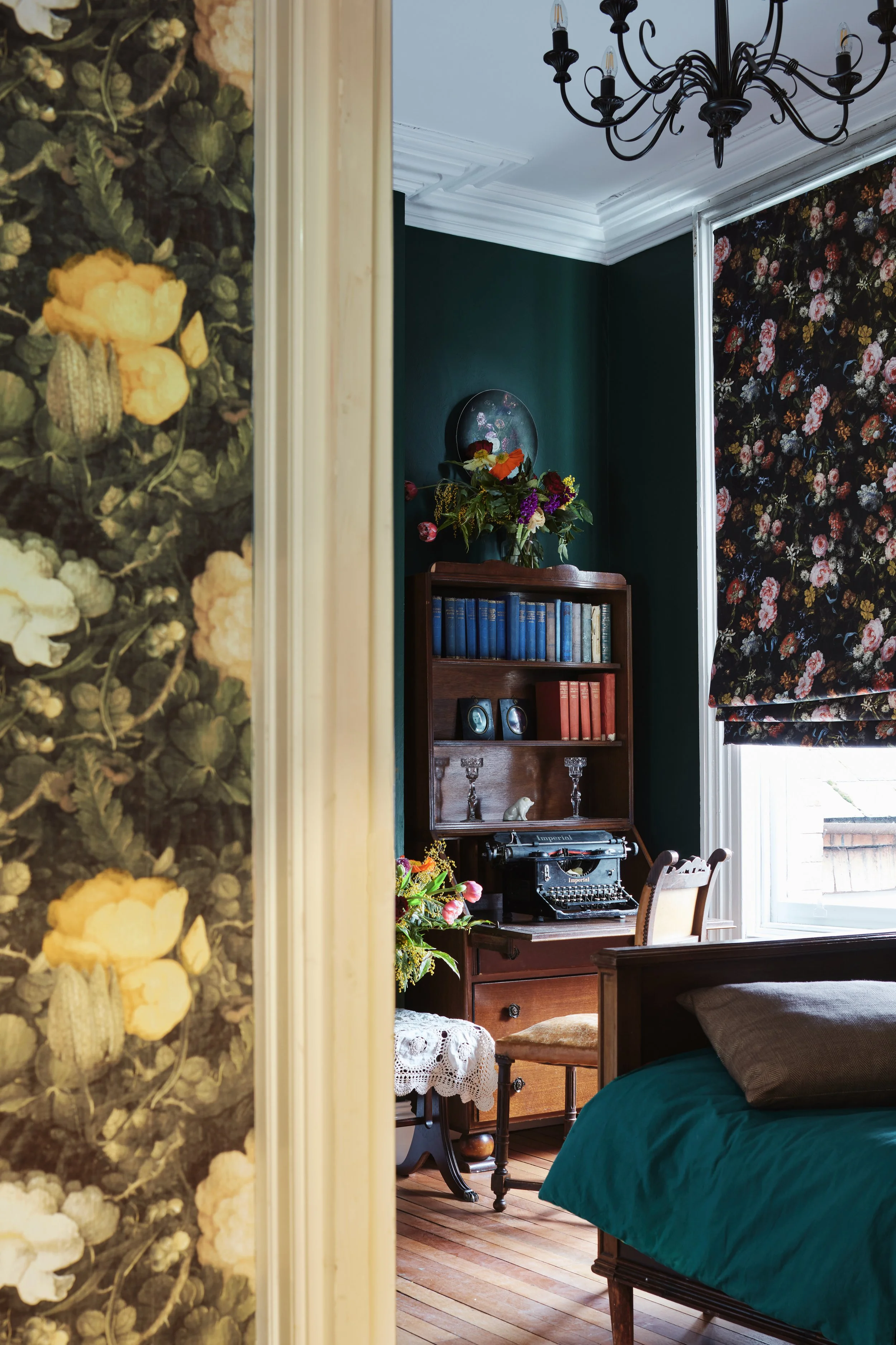 Vintage-inspired room with floral wallpaper, dark green walls, antique wooden desk, typewriter, floral decorations, bookshelf, and a decorative chandelier.