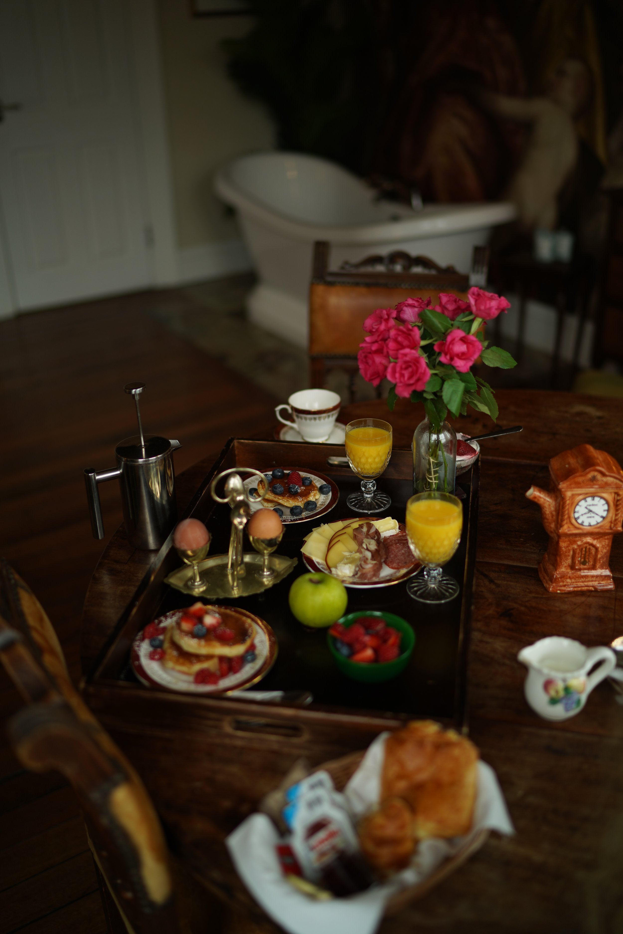 Breakfast setting with pancakes, eggs, cheese, salami, fruit, flowers, and orange juice on a wooden table, beside a vintage clock and a French press coffee maker.