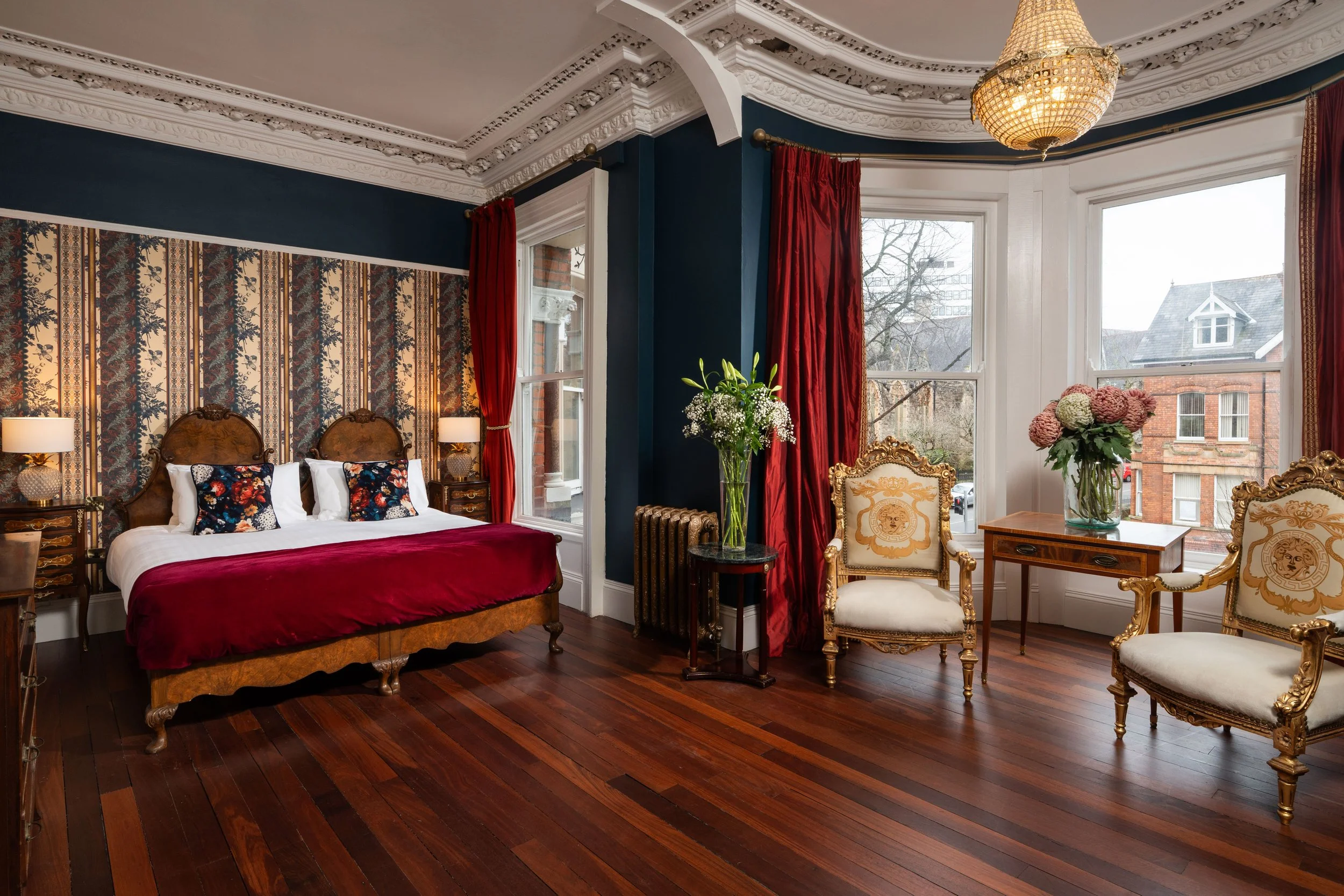 Luxurious bedroom with ornate furniture, large bed with red blanket, floral pillows, elegant chairs, wood flooring, chandeliers, and large windows with red curtains.